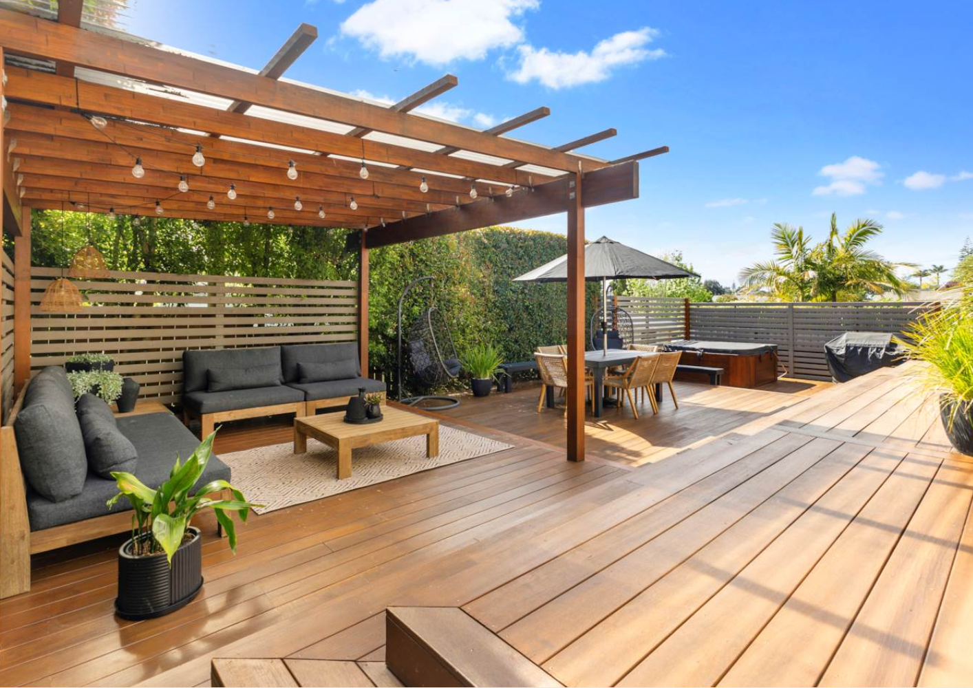 Spacious wooden rooftop terrace with outdoor seating, dining area, hot tub, and lush greenery under a blue sky with scattered clouds.