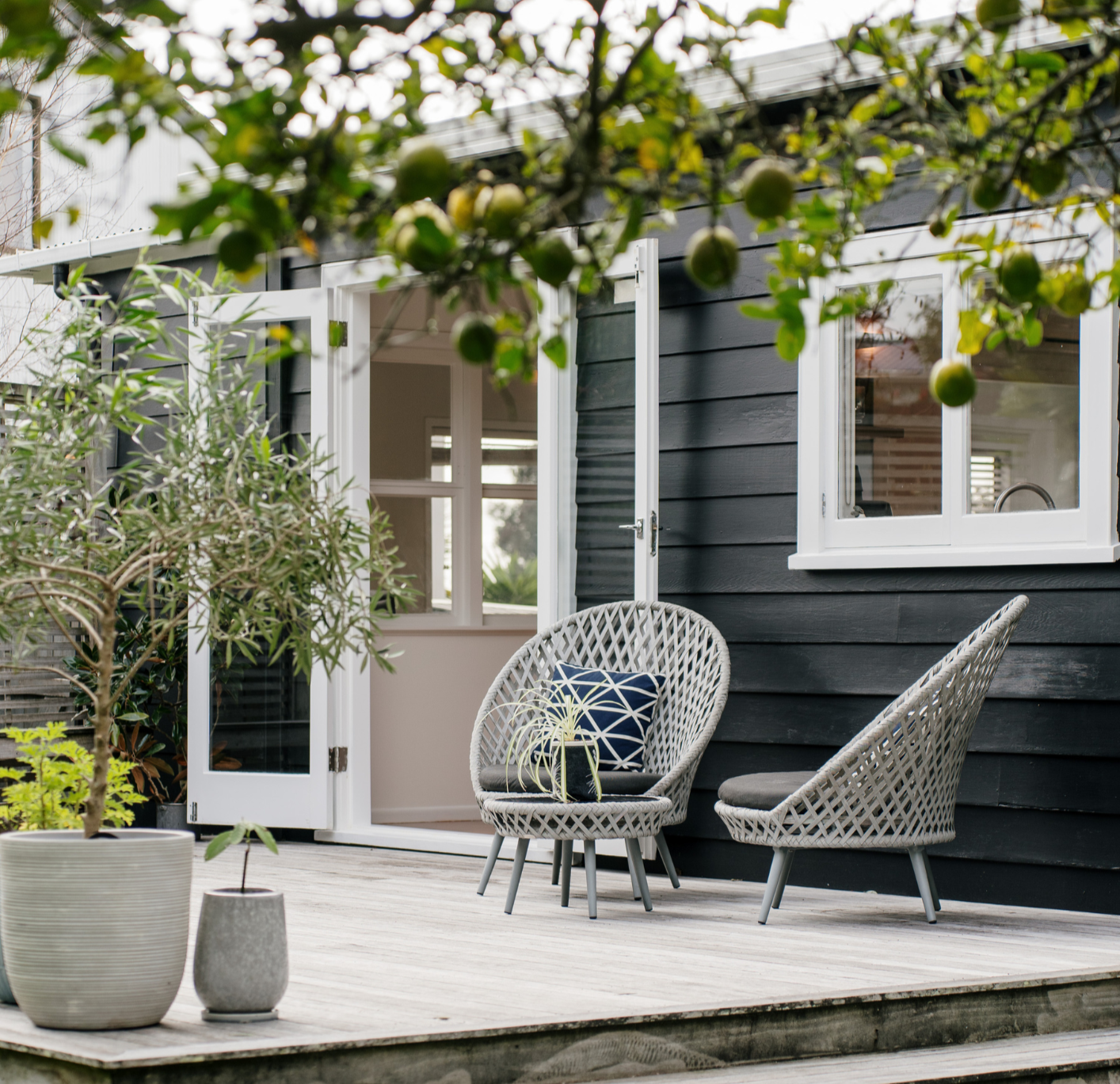 A cozy outdoor deck with two woven chairs, a small matching table with a plant, and potted greenery, attached to a modern black house with open white-framed windows and a door, with a tree with green fruit or flowers in the foreground.
