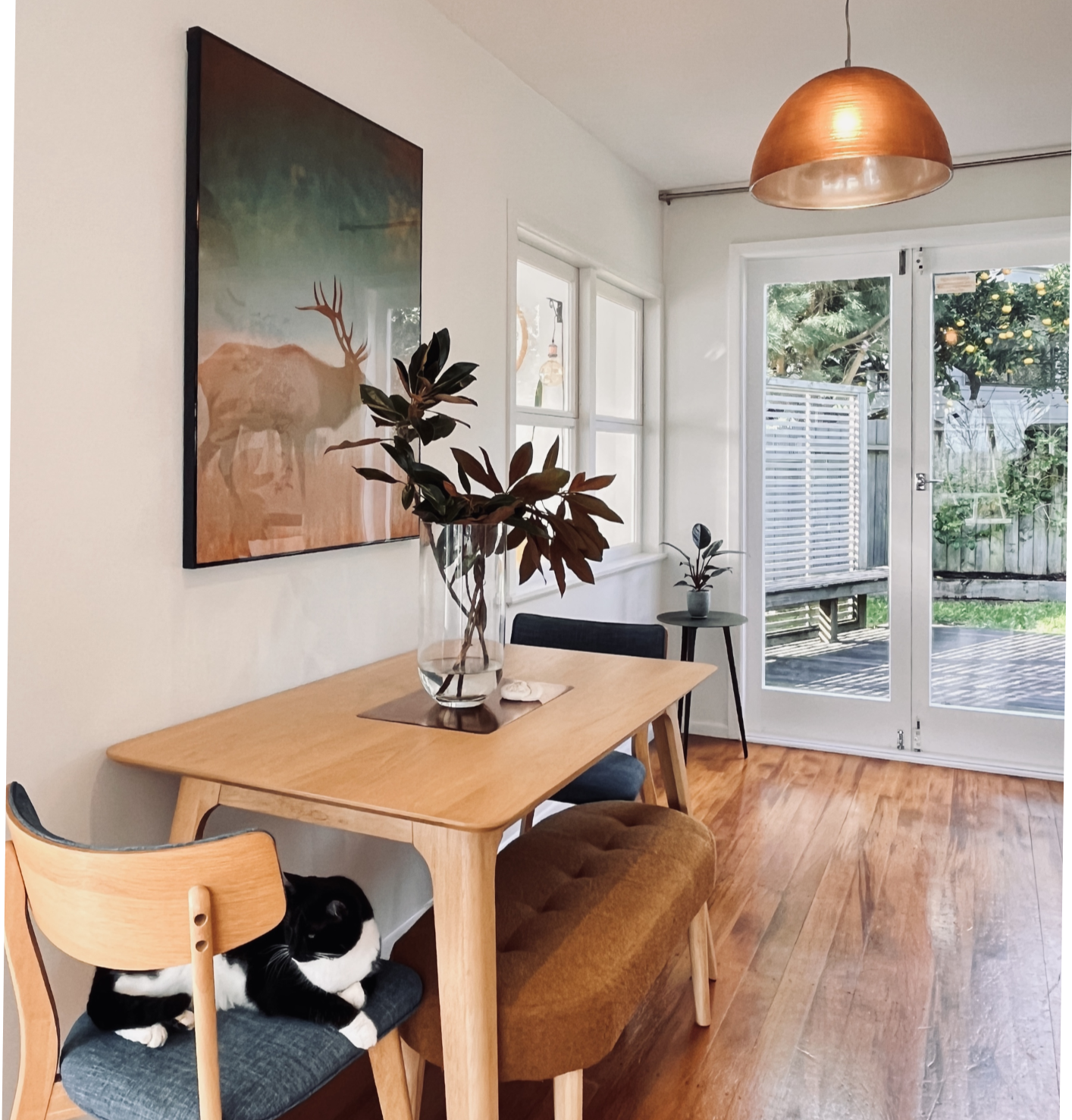 A cozy dining area with a wooden table, a glass vase with dark green leaves, a black and white cat lying on a chair, a large framed painting on the wall, and a sliding glass door leading to an outdoor patio with greenery.