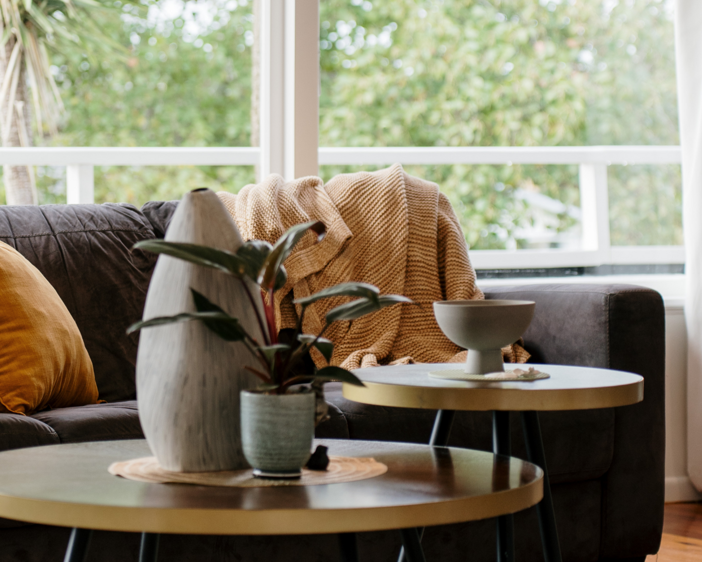 Living room with a dark sofa, a yellow pillow, a beige throw blanket, a round white side table with a beige bowl, another round side table with a large wooden sculpture and potted plant, and a window with green trees outside.