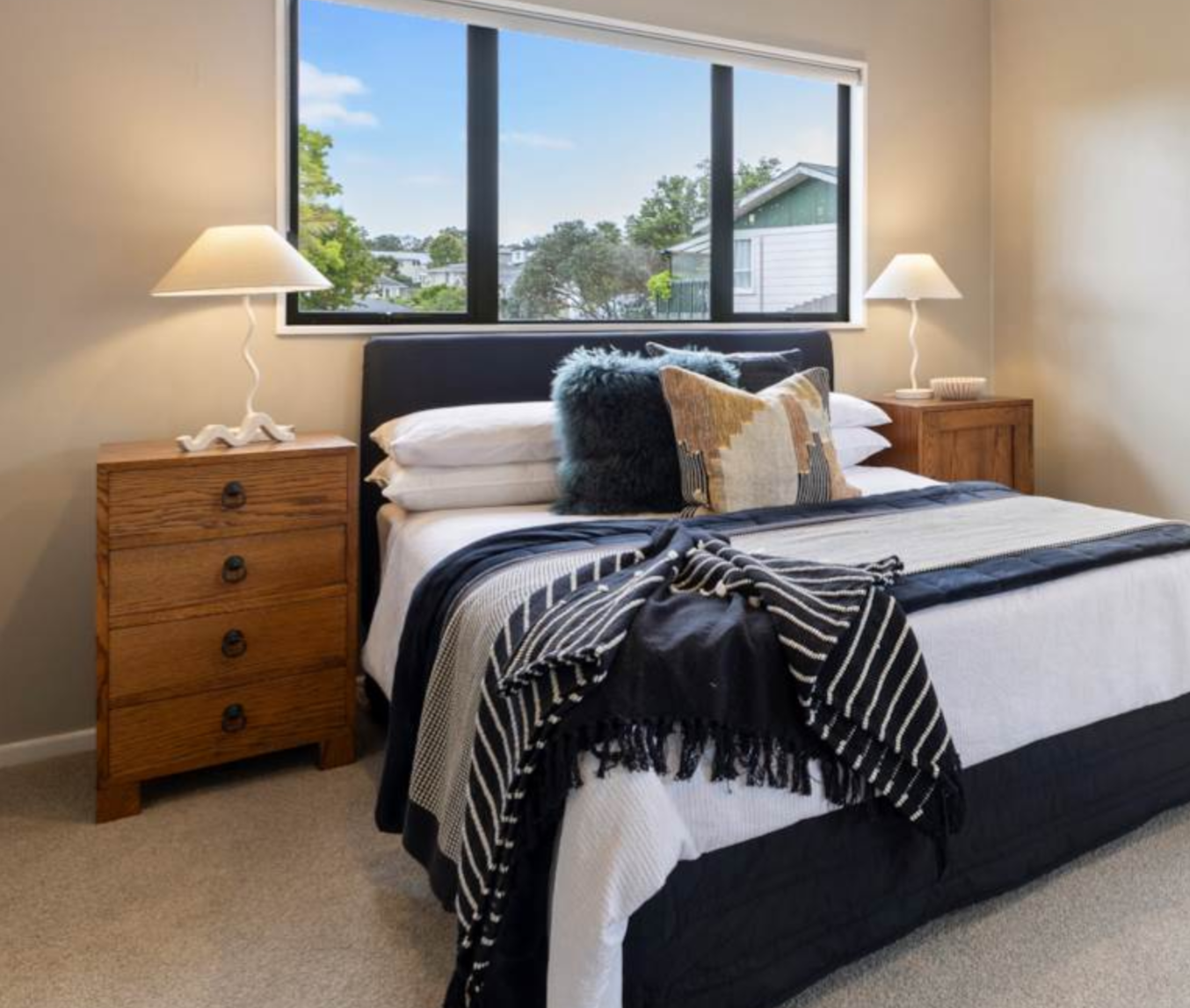 Bedroom with a bed, two wooden nightstands with white lamps, a window showing a neighborhood view, and a decorative pillow with a black and gold pattern.