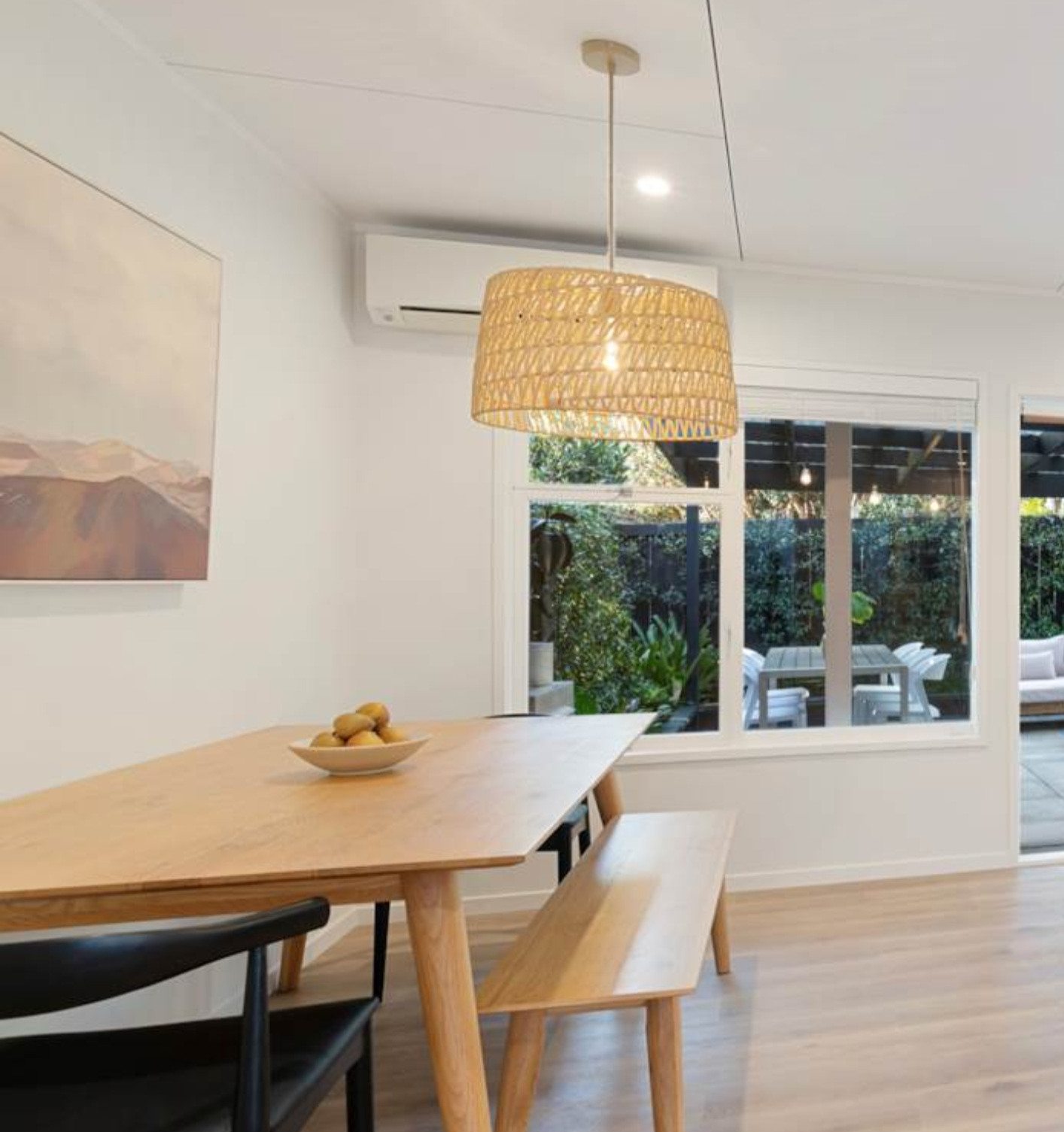 Interior of a dining room with a wooden table, a bench, a black chair, a bowl of lemons, a hanging wicker light fixture, and large windows with view of outdoor patio and greenery.