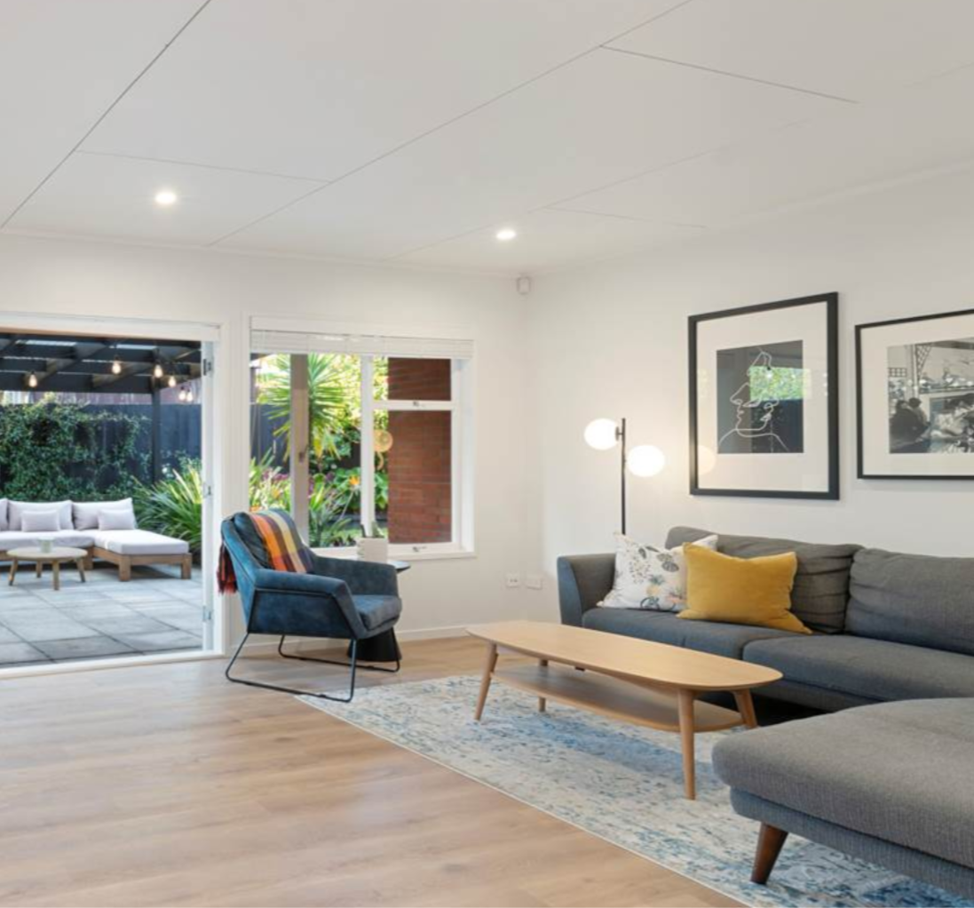 Modern living room with a grey couch, armchair, and wooden coffee table, artwork on the wall, and a view of a patio with outdoor seating and greenery