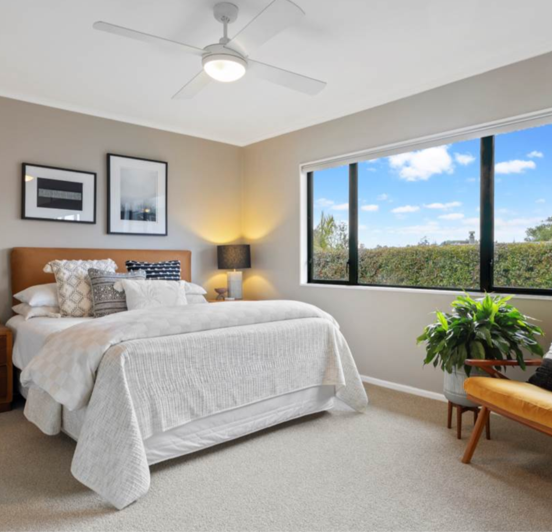 Bedroom with a bed, pillows, a nightstand with a lamp, a window showing trees and blue sky, a chair with a yellow cushion, and a large green plant.