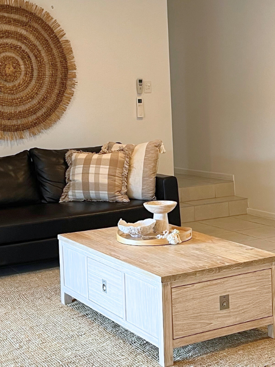 Living room with a black leather sofa with plaid and solid pillows, a beige and white wooden coffee table with decorative bowls, a round woven wall hanging, and a stairway in the background.