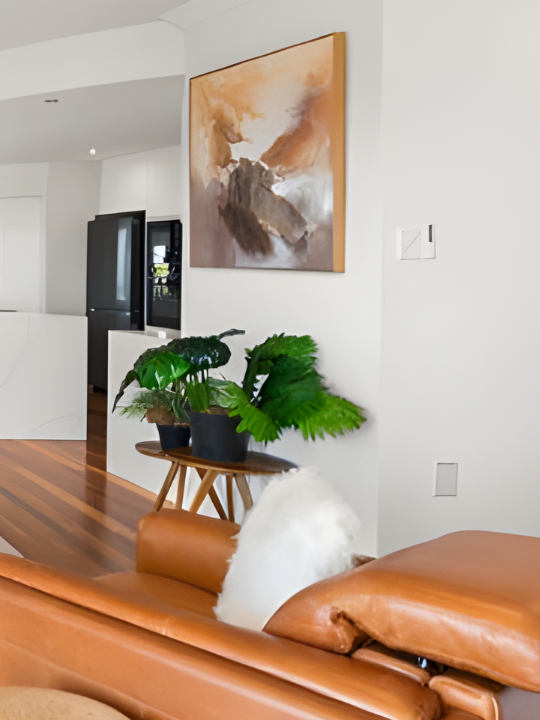 Interior of a modern living room with a leather couch, a plant on a small wooden table, abstract wall art, and hardwood floors.