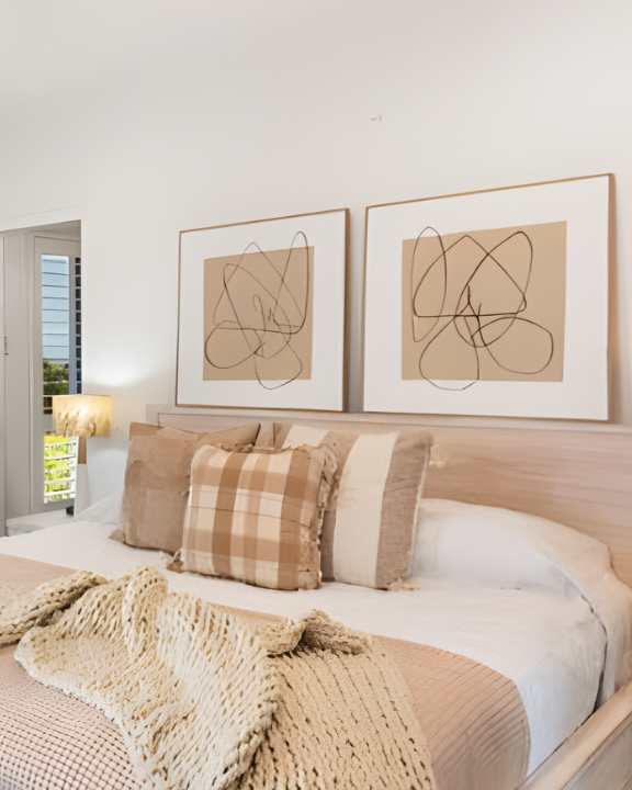 A bedroom with a bed, beige pillows, a knitted blanket, and abstract art prints hanging above the headboard.
