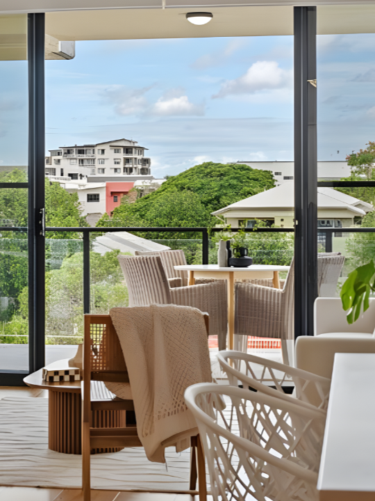View from a modern apartment balcony with outdoor chairs and a table, overlooking trees and neighboring buildings under a partly cloudy sky.