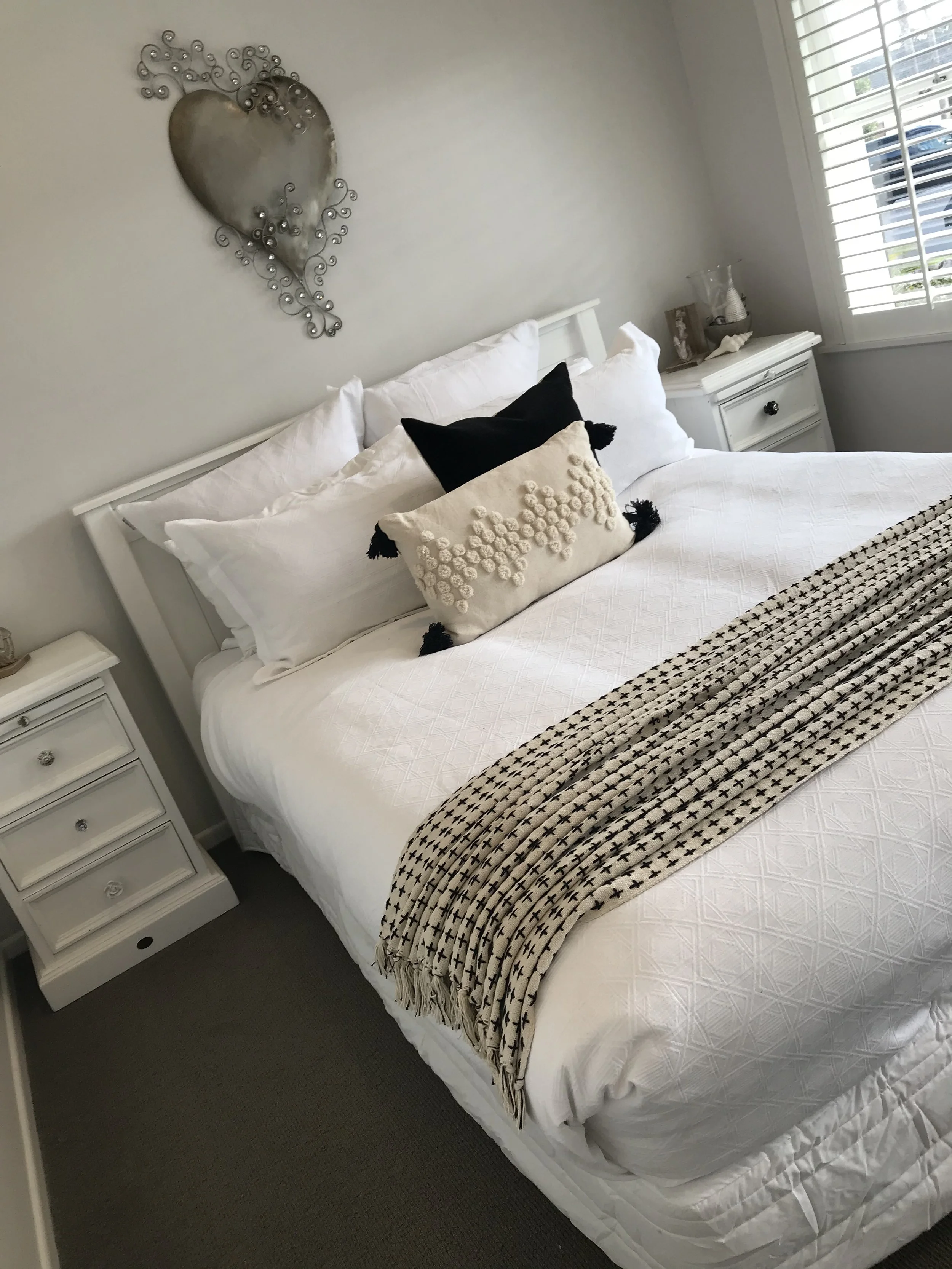 A neatly made bed with white sheets and pillows, a decorative black and beige throw blanket, and a textured beige decorative pillow with tassels. There are two white nightstands, one on each side of the bed, with a metal heart wall decor above the he