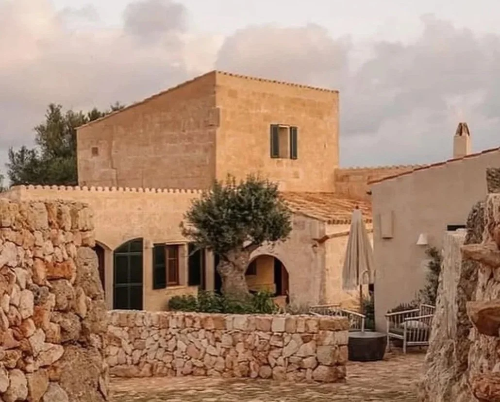 A stone building with a ladder on the roof, a small arched doorway, and windows. There is a large tree in front, stone walls, and patio furniture with an umbrella. The setting appears rustic and Mediterranean.