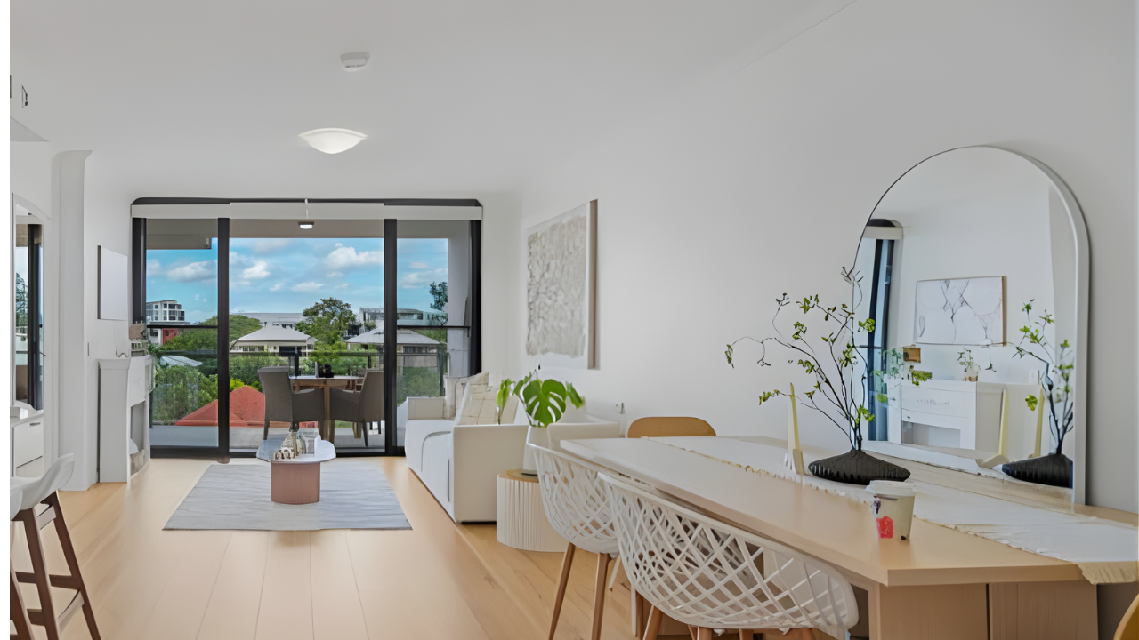 Bright living room with white walls and light wood flooring, featuring a white sofa, a small dining table with chairs, a large mirror, and a sliding glass door opening to a balcony with outdoor seating and a view of houses and trees.