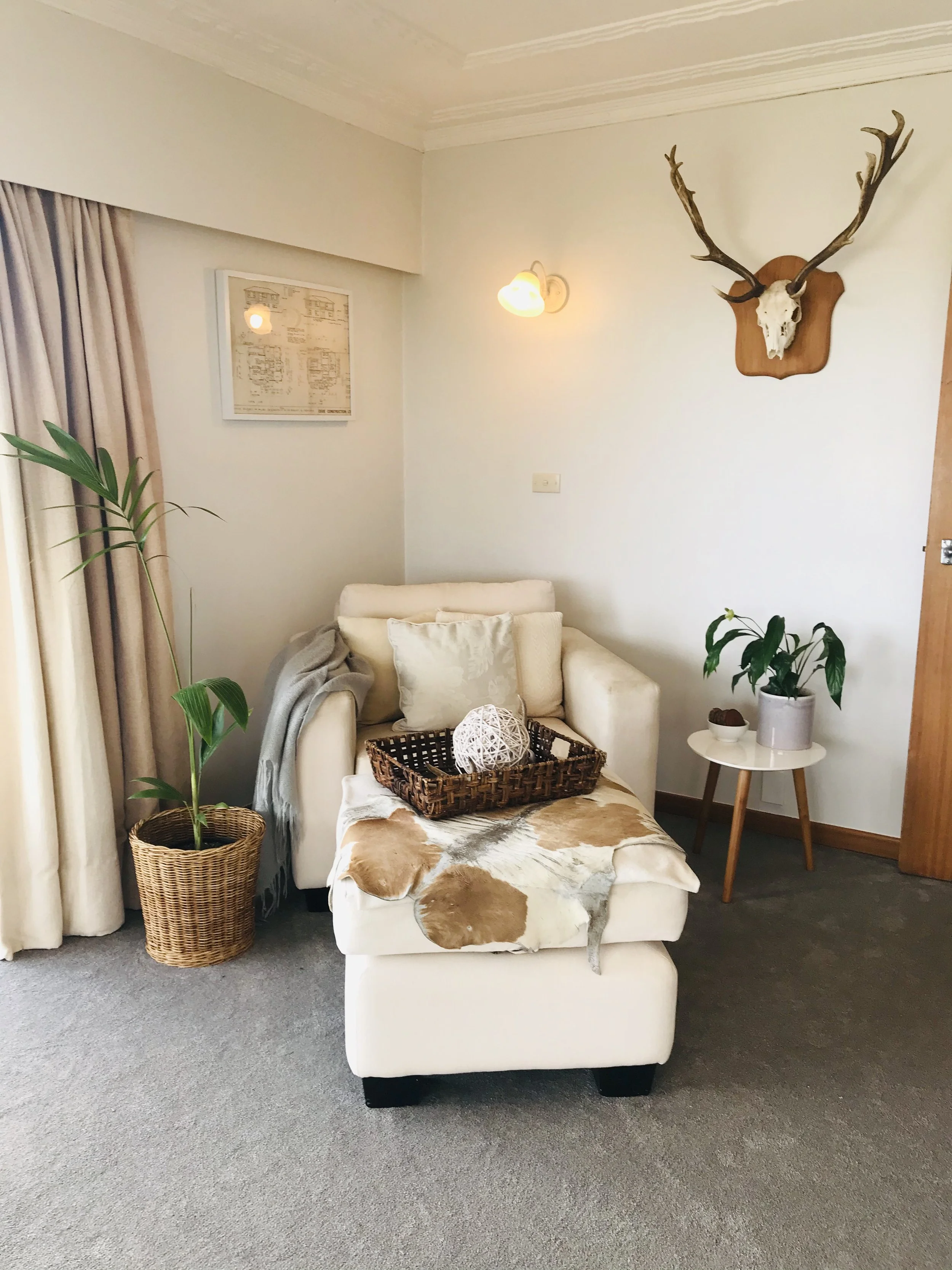 A cozy living room corner with a white sofa, striped cowhide ottoman, and potted plants. Decor includes a mounted deer skull with antlers on the wall, a framed picture, a wall lamp, and a small table with a potted plant and decorative items.