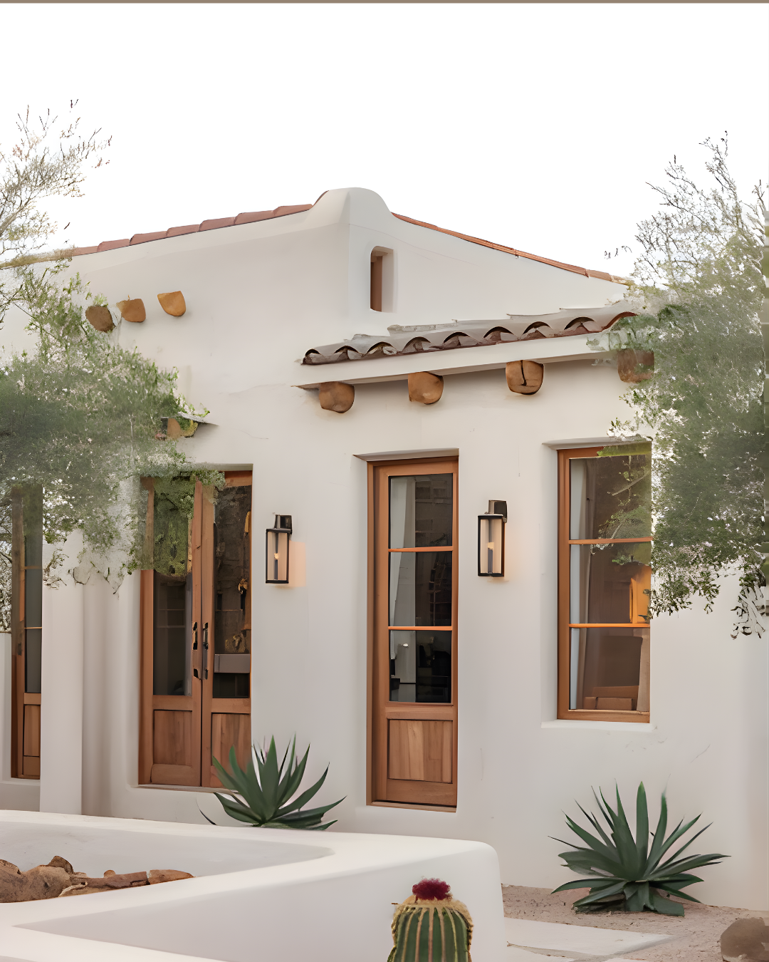 A white stucco house with wooden window frames, desert plants, and a small cactus in the foreground, with a desert landscape and trees around.