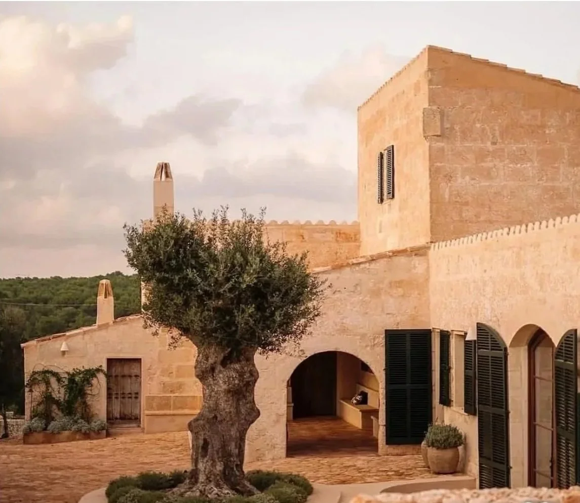 A Mediterranean-style stone building with black shutters, a courtyard, and an olive tree in the foreground.