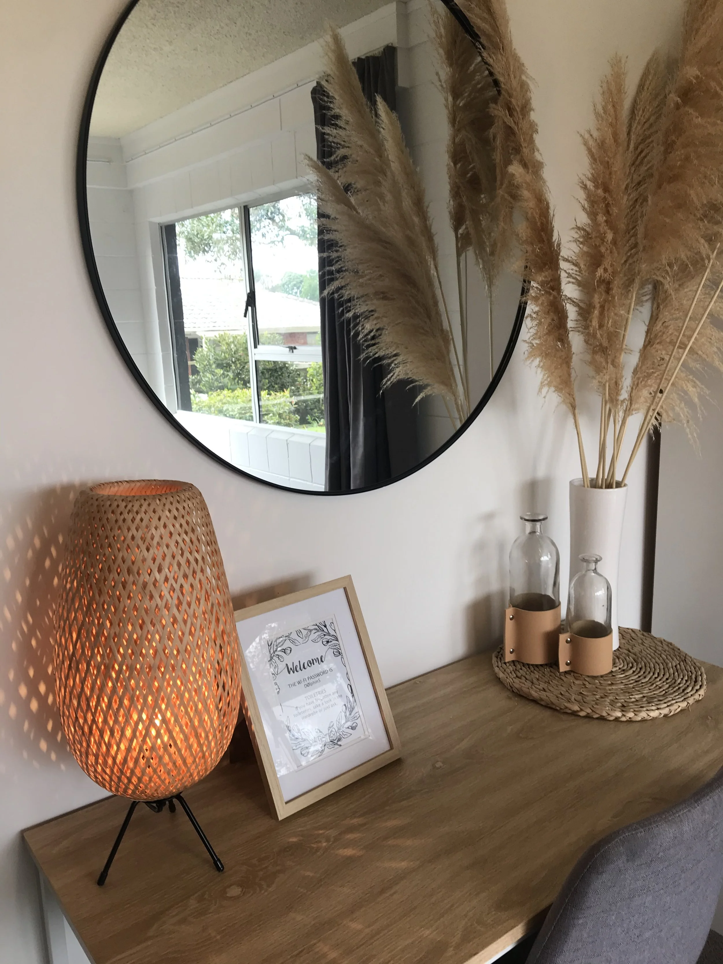 Decorative wooden table with a woven lamp, framed welcome sign, glass bottles, and a tall vase with pampas grass, reflected in a round wall mirror.
