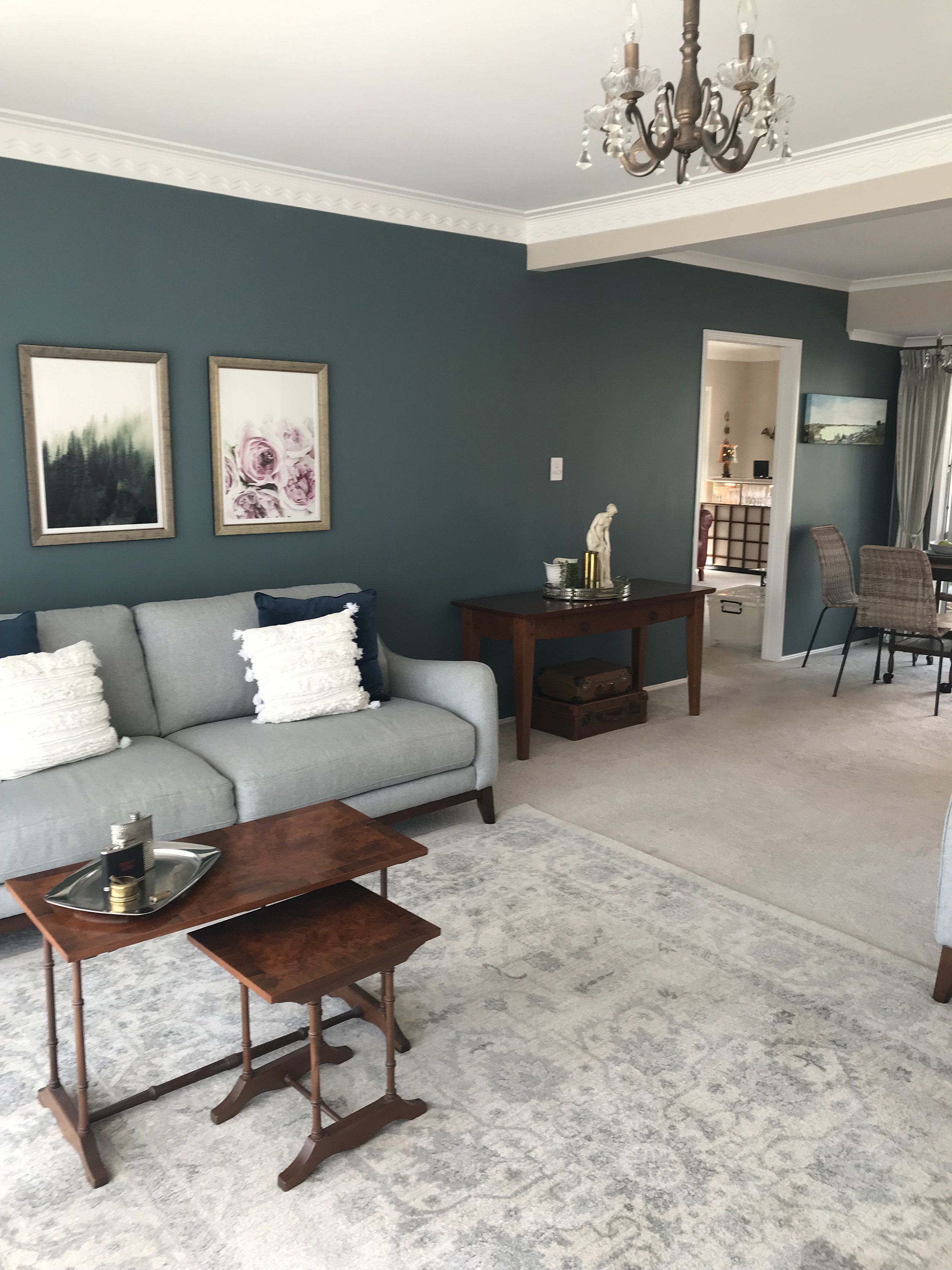 Living room with gray sofa, wooden tables, and wall art, adjacent to dining area with curtains, chandelier, and decorative items.