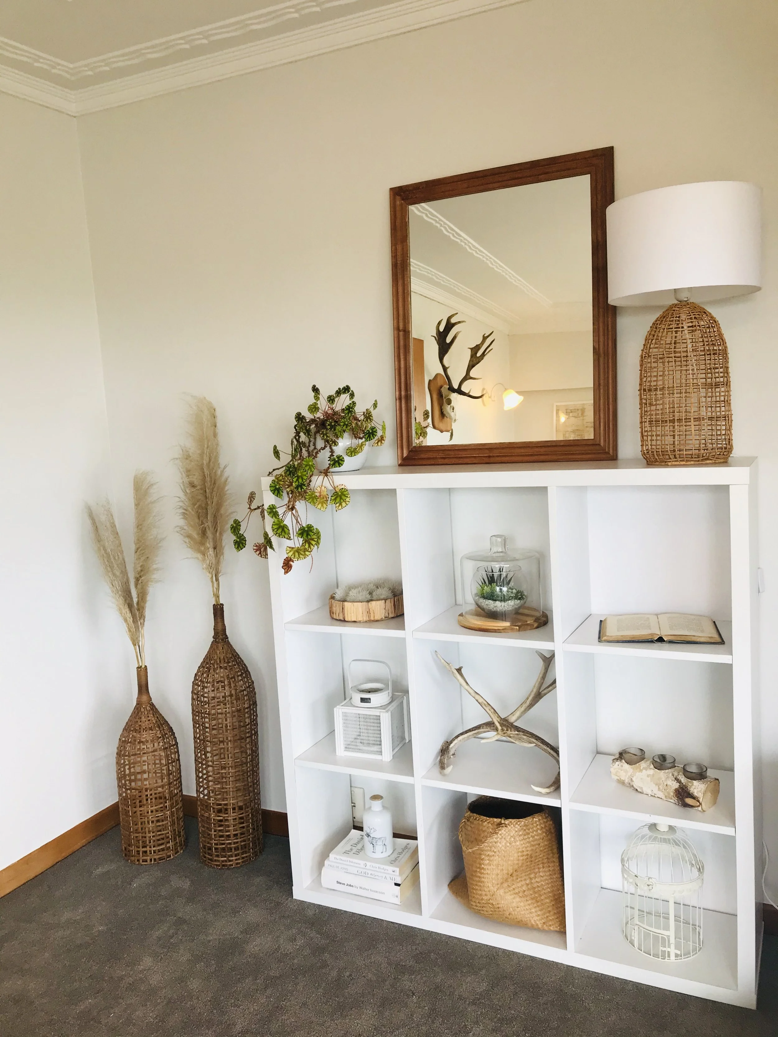 Decorative white bookshelf with glass terrarium, woven basket, antlers, open book, candles, and potted plant, next to two tall wicker vases with pampas grass, mirror with wooden frame, and a wicker lamp on top of the shelf in a living room corner.