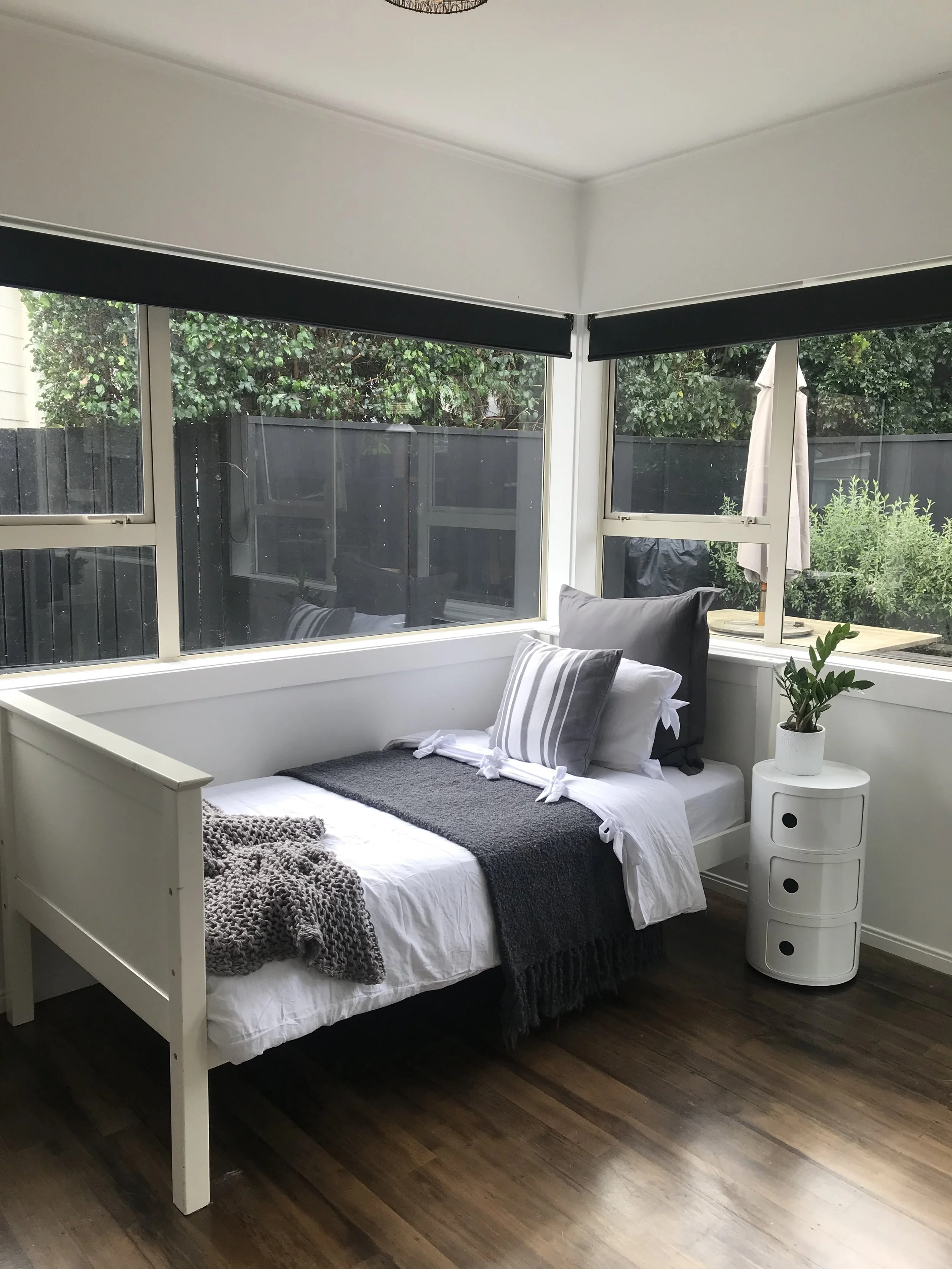 Bedroom with large windows, white bedframe, gray blanket, and a small white curved nightstand with a potted plant, hardwood floor.