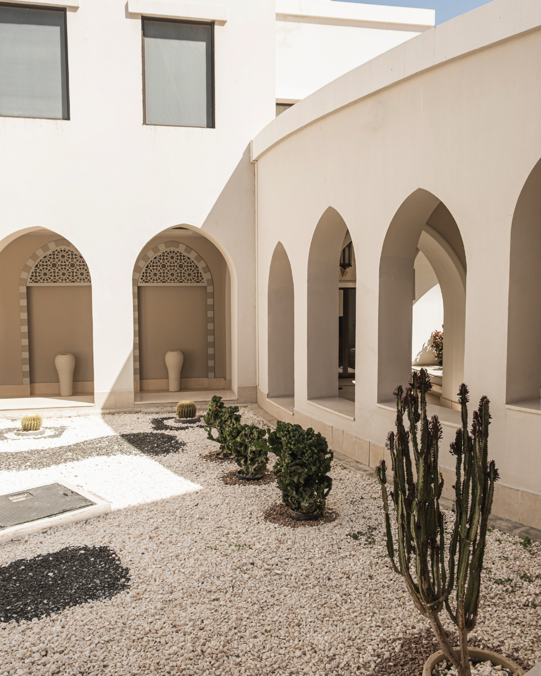 Exterior view of a white Mediterranean-style building with arched doorways and windows, decorative urns, cacti, and gravel landscaping.