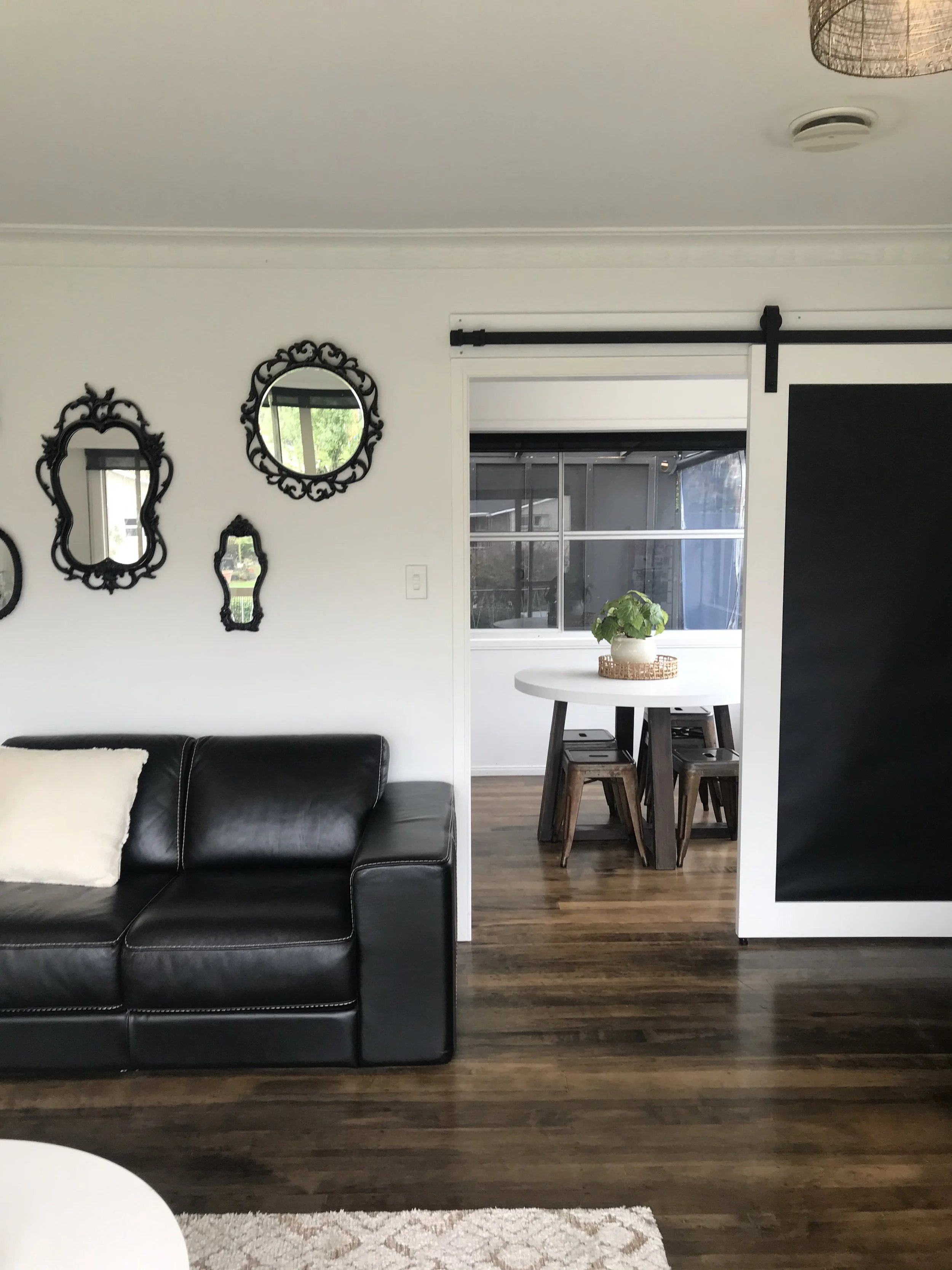 Living room with black leather sofa, white pillow, and wall with decorative black mirrors. Sliding barn door leads to dining area with wooden table, stools, and a potted plant.
