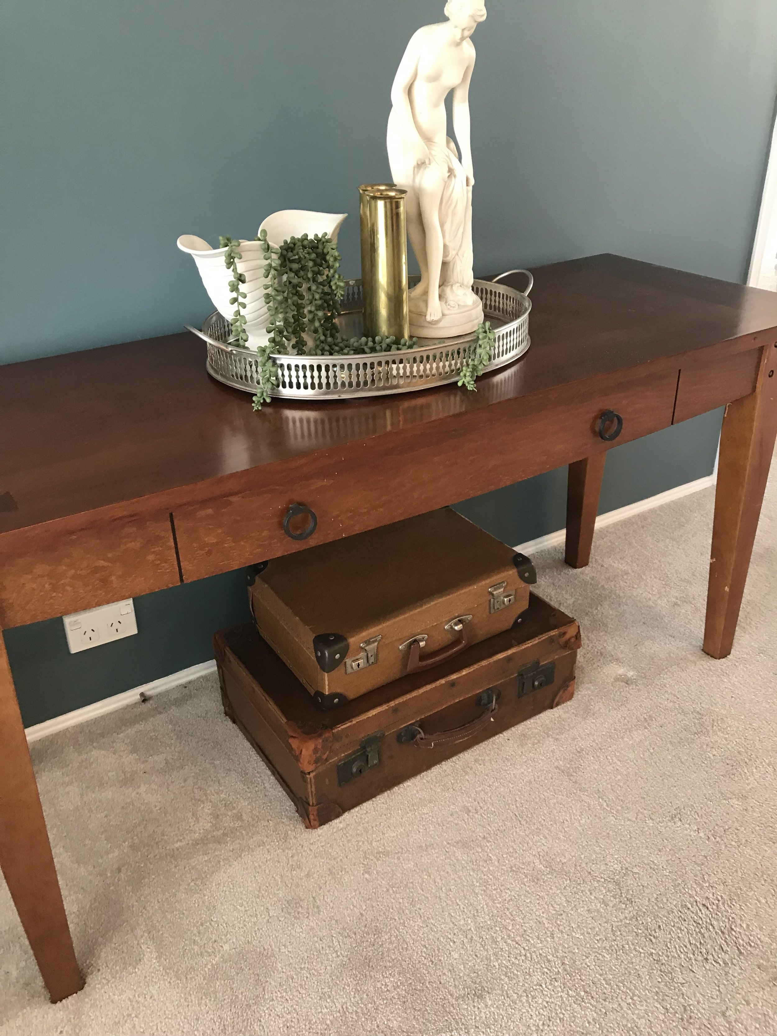A wooden console table decorated with a silver tray holding a white pitcher with a green trailing plant, a pair of gold-colored vases, and a white statue of a woman. Two stacked vintage suitcases are underneath the table.