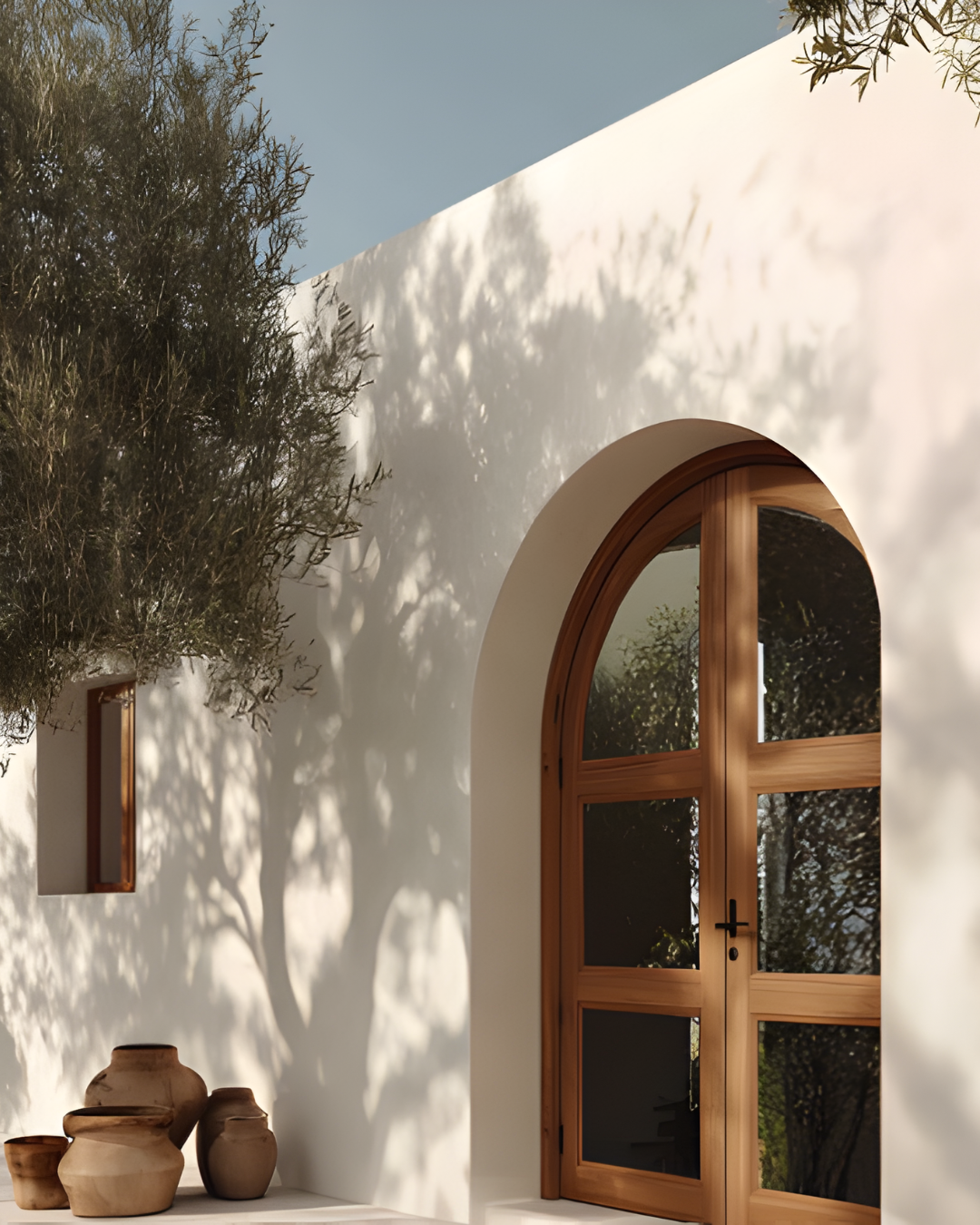White building with arched wooden window, small rectangular window, and pottery vases outside, with trees casting shadows on the wall inside a sunny outdoor setting.