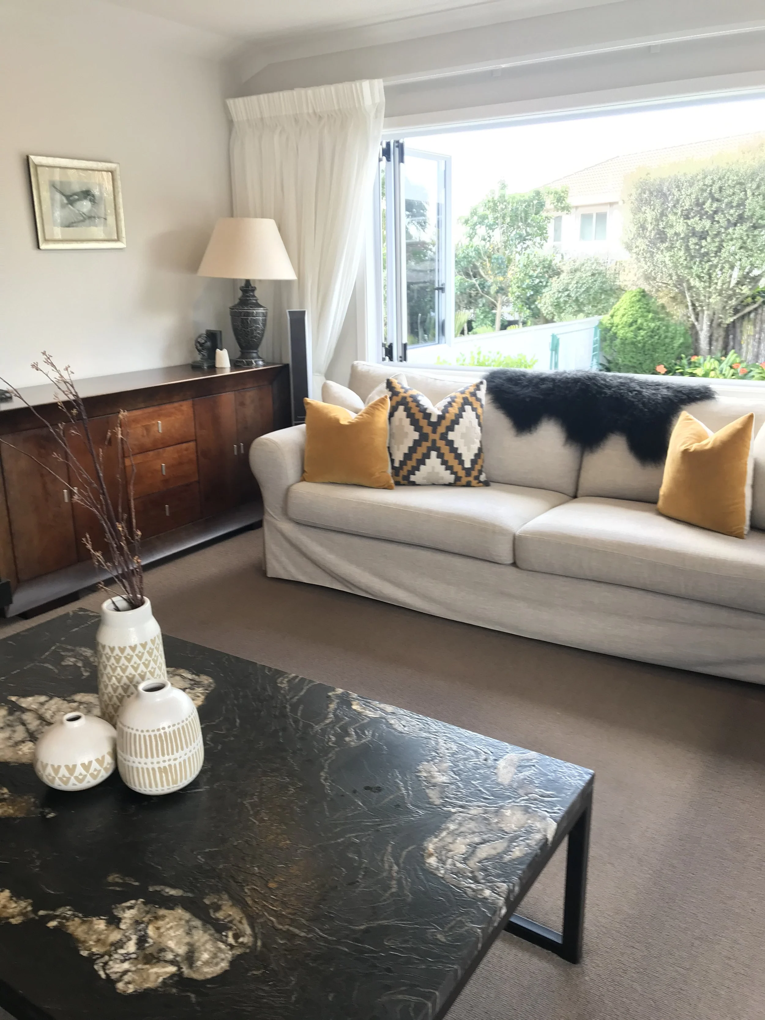 Living room with beige sofa, four decorative pillows, a dark wooden sideboard with a lamp, a picture frame, a tall black speaker, an open window showing greenery outside, and a black marble coffee table with three white vases.