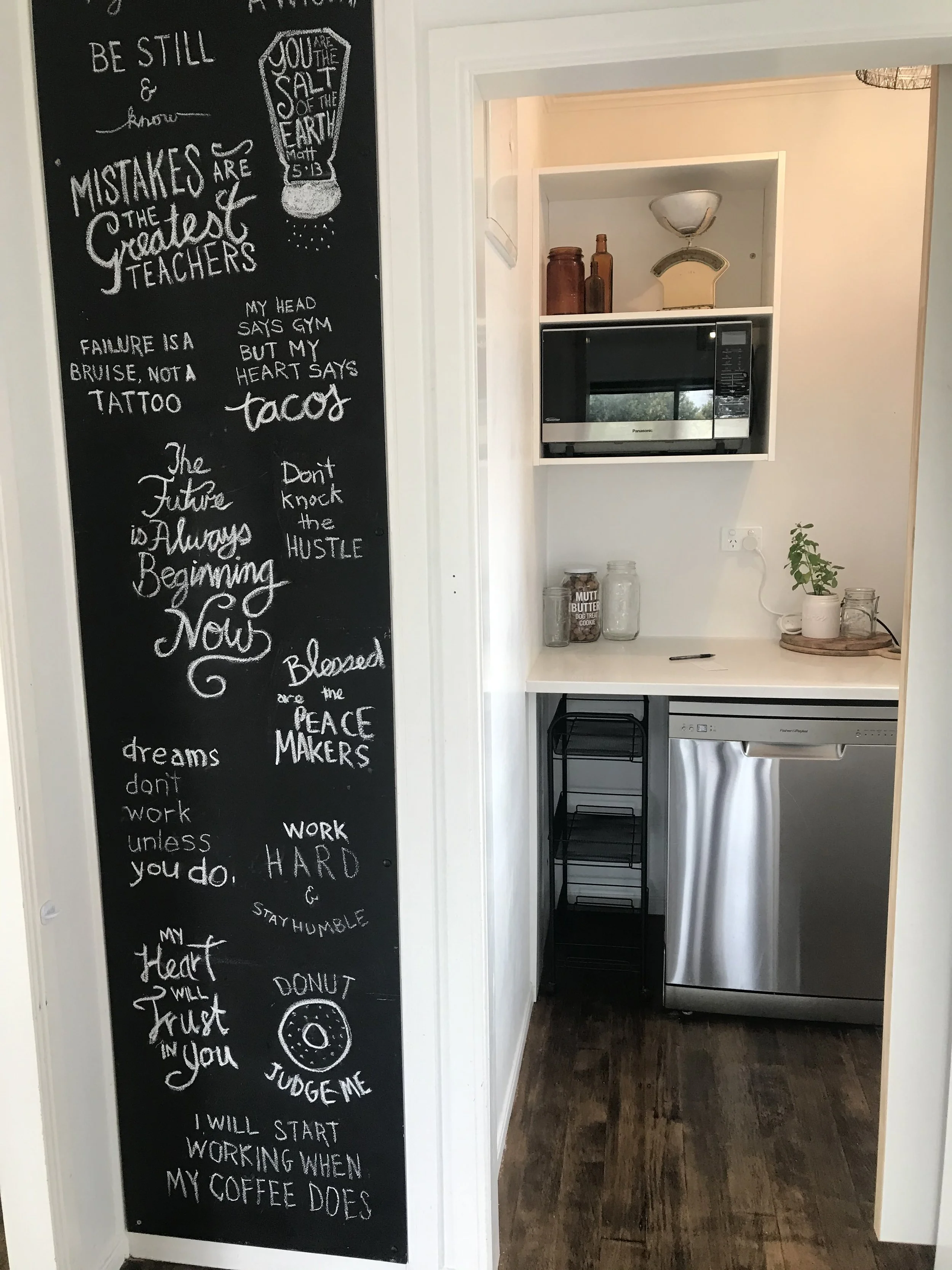 A black chalkboard wall with various handwritten quotes and messages in white chalk, adjacent to a small kitchen nook with a microwave, jars, a plant, and a dishwasher.