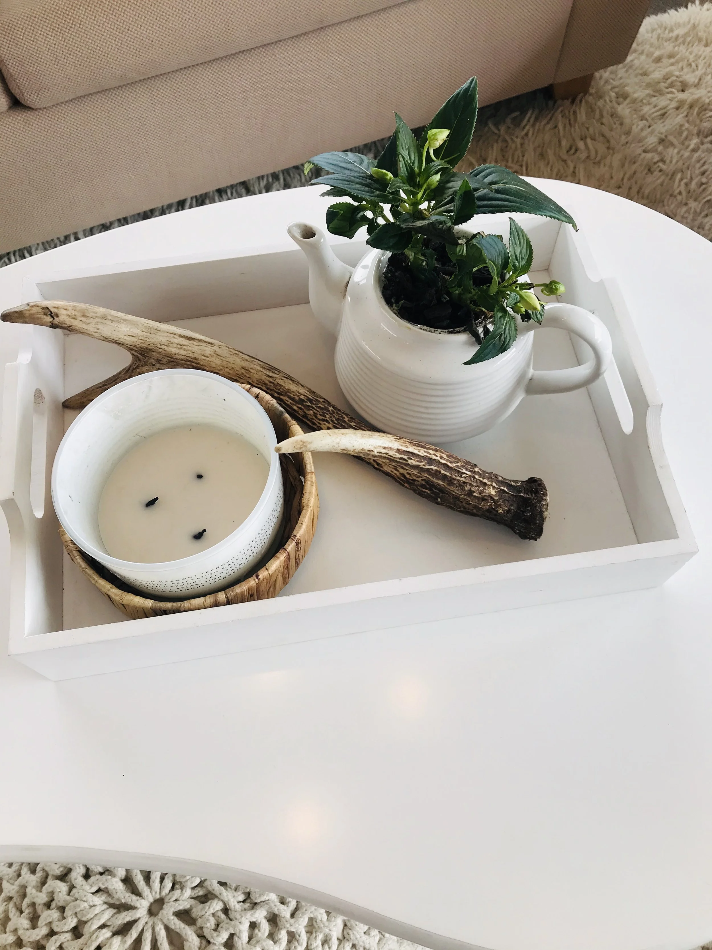 Decorative tray with a white teapot holding a green plant, a burning white candle, and two brown sticks, placed on a white table.