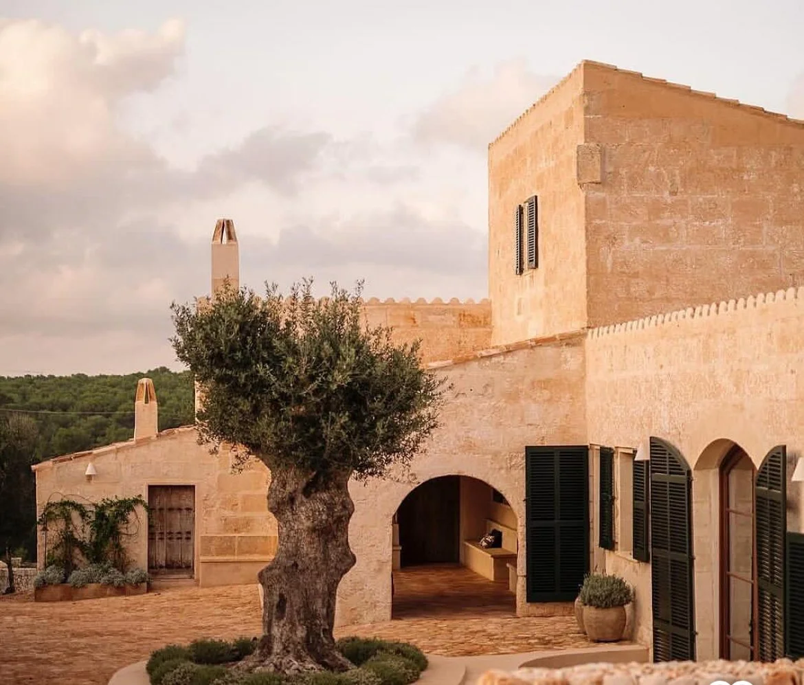 A sunlit stucco house with black shutters, a small tree in a planter, and a cobblestone courtyard.