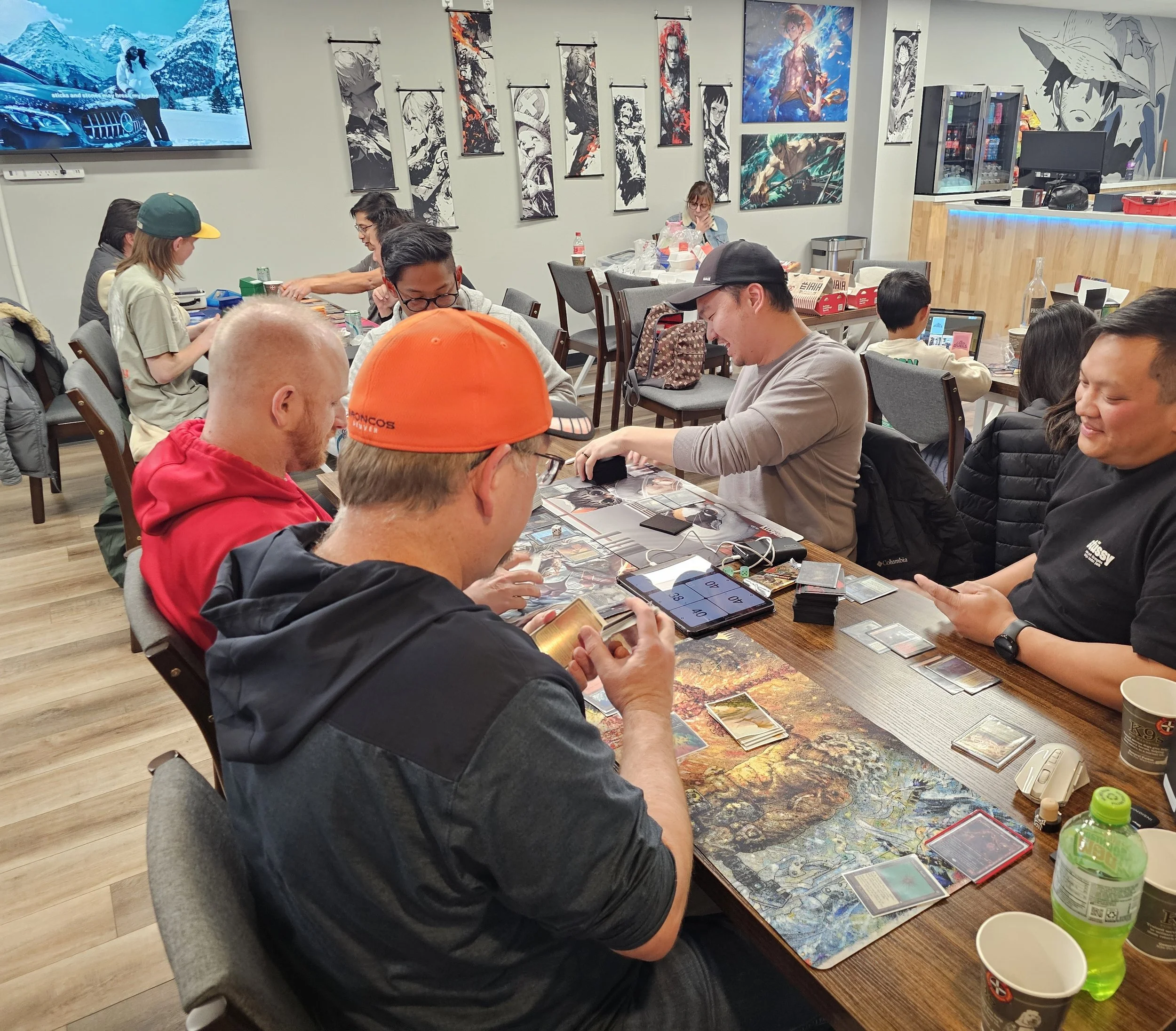 People playing tabletop card and strategy games at a long wooden table in a gaming cafe or community center, with artwork and posters on the walls, screens showing digital content, and drinks on the table.