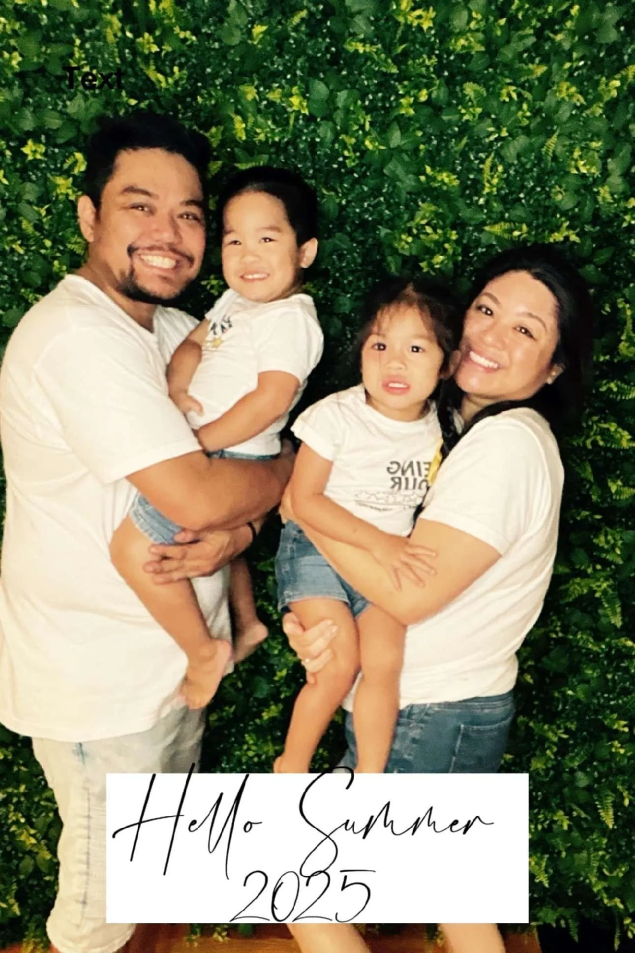 A happy family of four, two adults and two children, smiling and holding each other in front of a lush green bush. The man is on the left, holding a girl with short black hair. The woman on the right is holding another girl with curly hair. Both children are wearing white T-shirts and denim shorts. There is text at the bottom that says, 'Hello Summer 2025'.