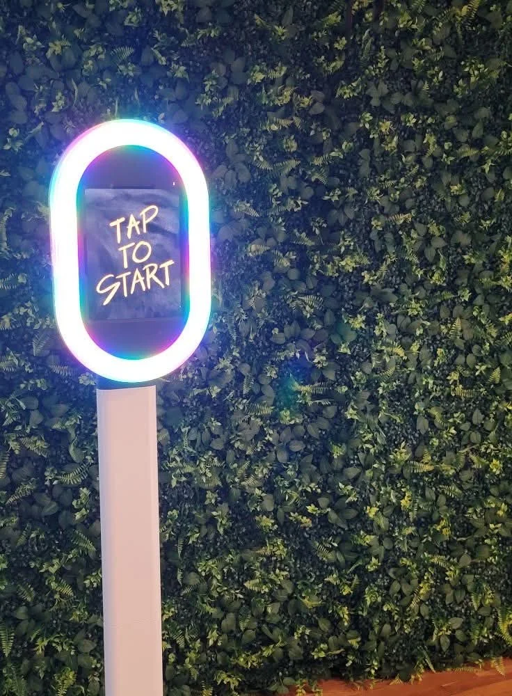 A neon-lit kiosk with a screen displaying the words 'Tap to Start' against a lush green leafy wall background.