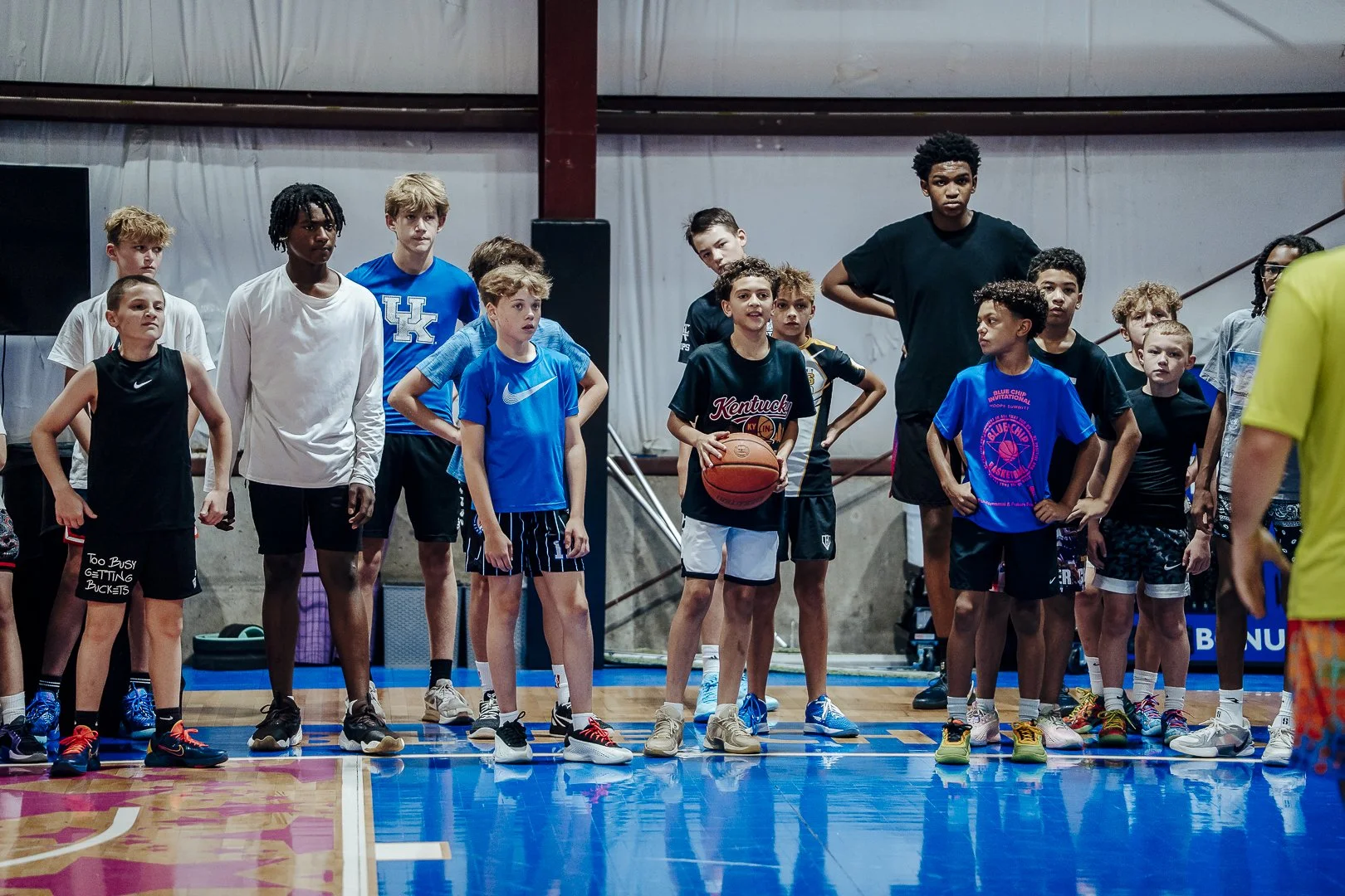 A group of young boys and a tall teenager, wearing basketball attire, standing on an indoor basketball court during a practice or team meeting. One boy near the center is holding a basketball, and others have hands on hips or are attentively listening.