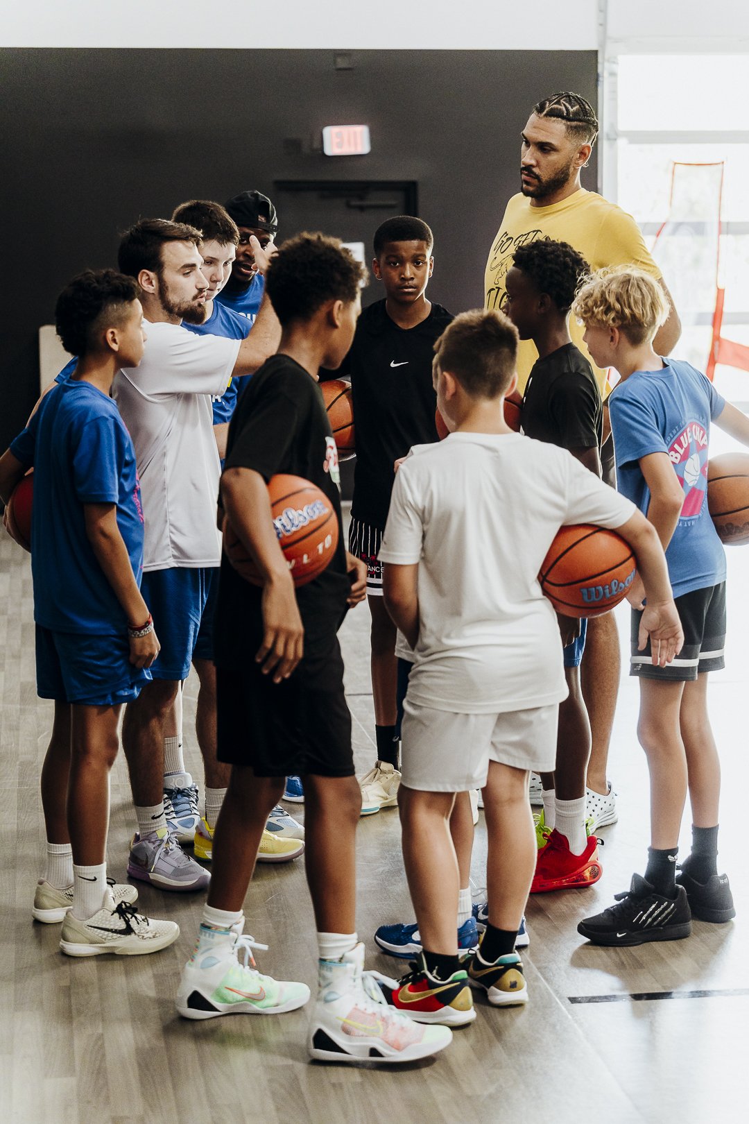 A group of young boys and adult males in a basketball gym, holding basketballs, gathered around a coach or instructor who seems to be giving instructions.