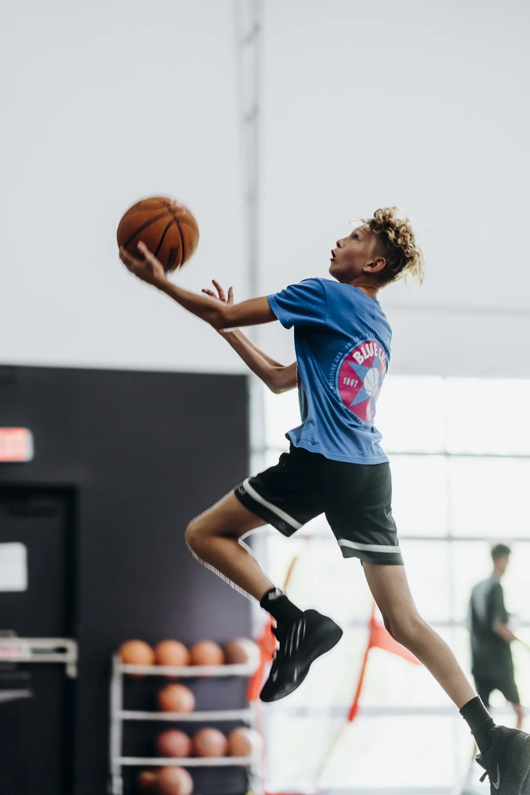 Young boy playing basketball indoors, jumping to shoot the ball, wearing a blue t-shirt, black shorts, and black shoes.