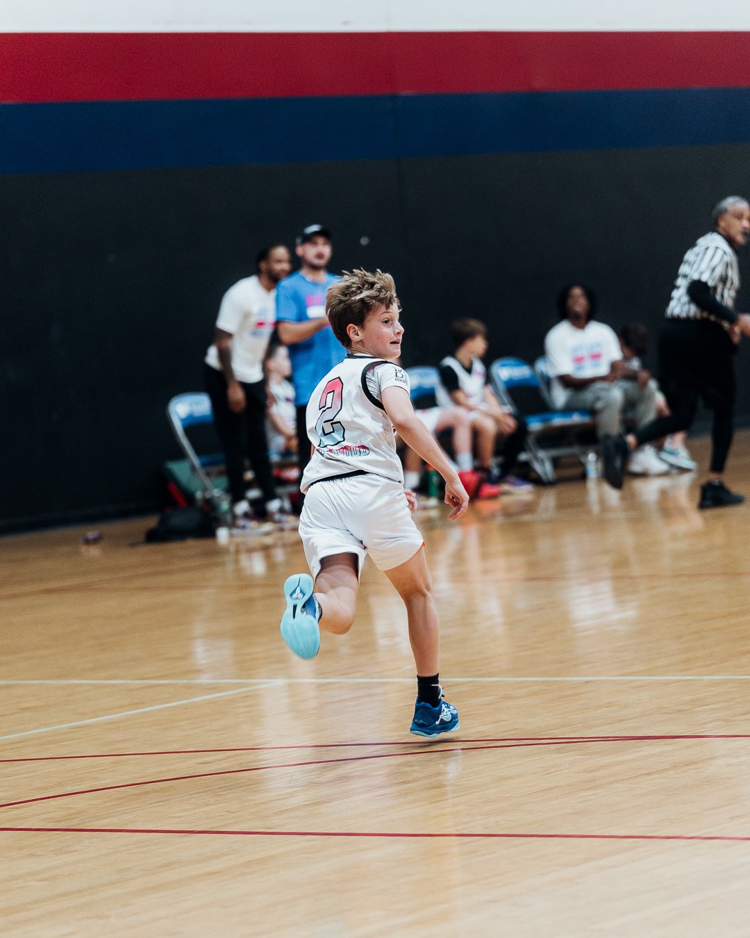 A young boy running on an indoor basketball court during a game, with teammates and a referee watching from the bench.