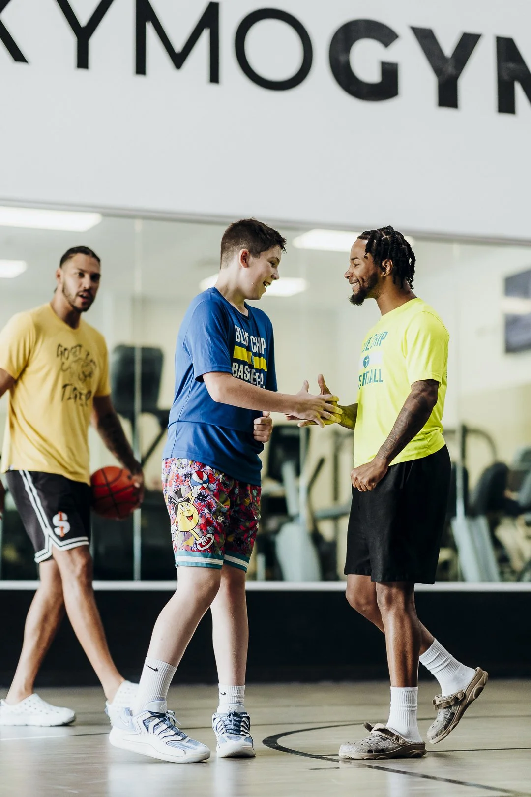 Two young men shaking hands on a basketball court while a third man holding a basketball watches. All are casually dressed for basketball practice or game in a gym with exercise equipment visible in the background.