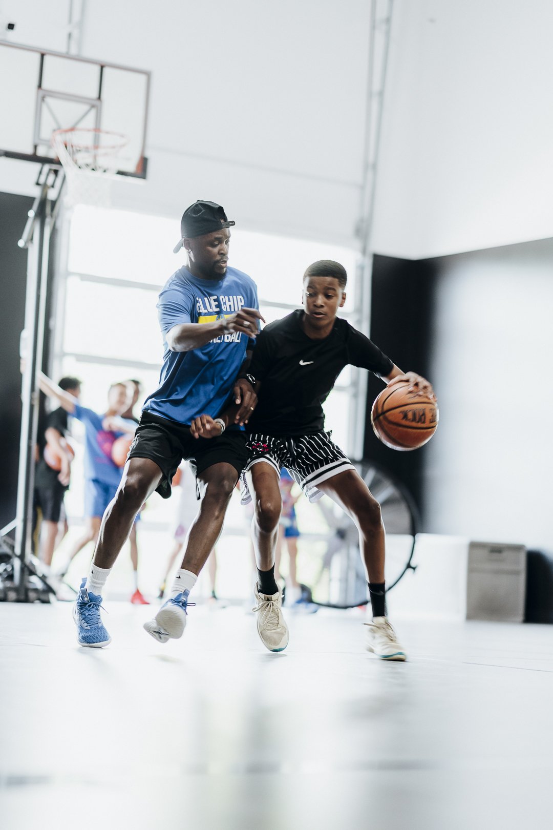 Two boys playing basketball indoors, one in a black shirt and striped shorts dribbling the ball while the other in a blue shirt and black shorts defends.