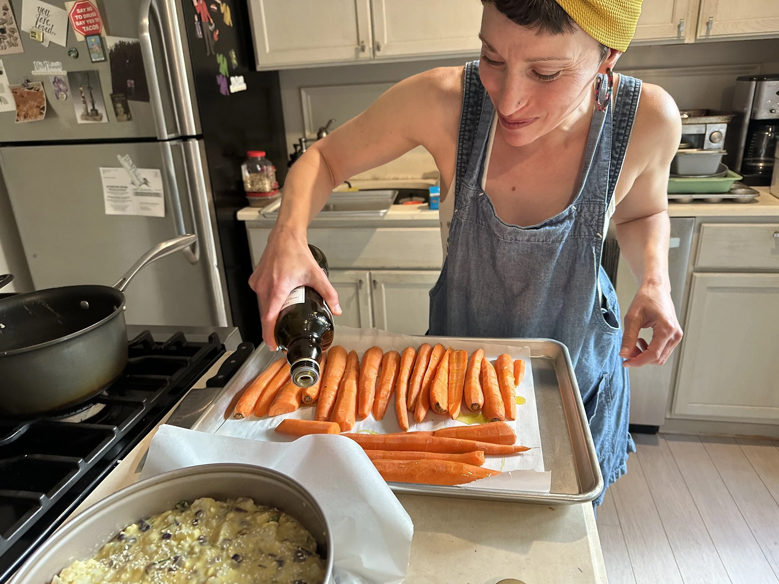A woman sprinkling seasoning on carrots arranged on a baking sheet in a kitchen.