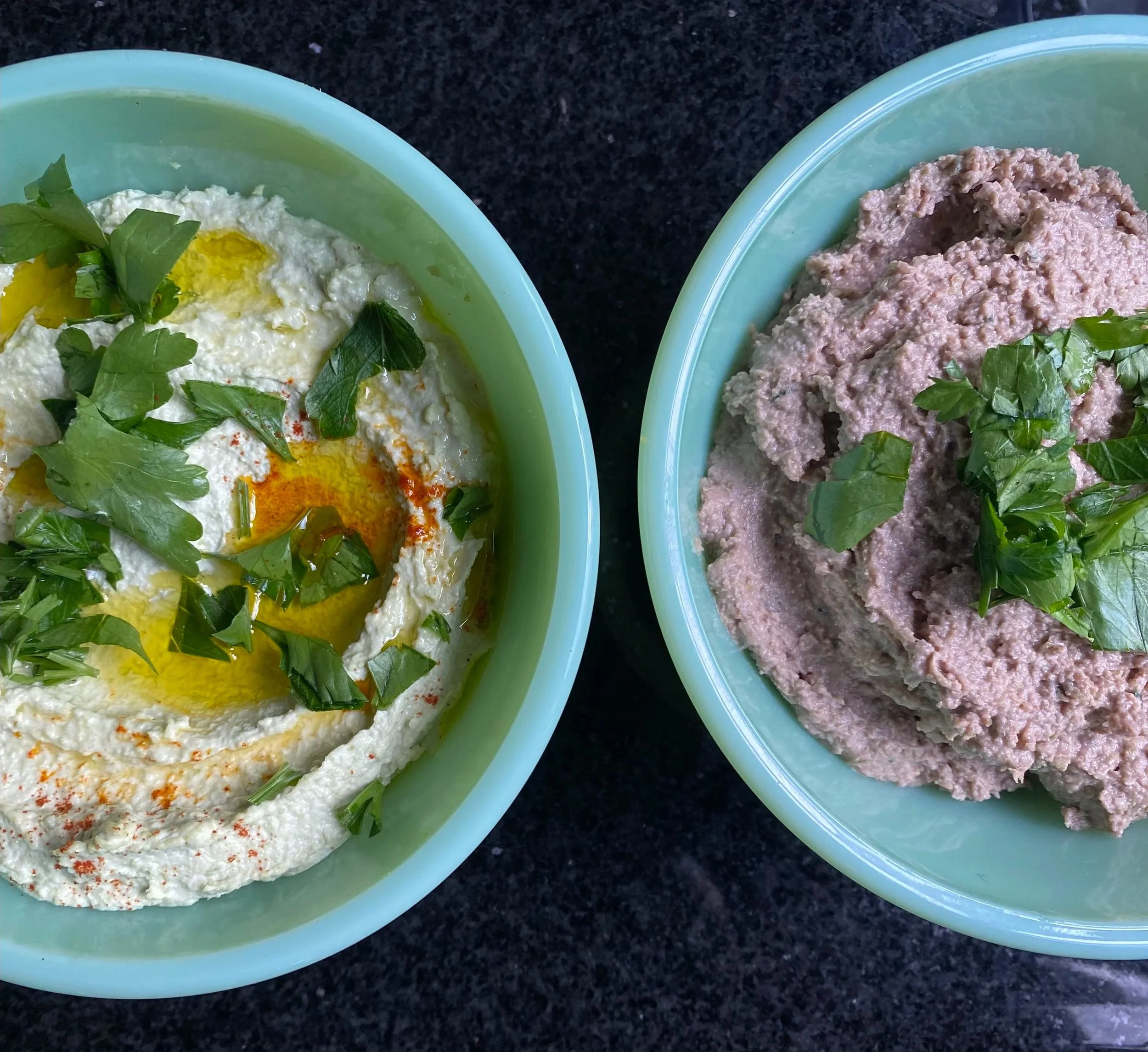 1 bowl of edamame hummus and lentil walnut pate garnished with chopped fresh herbs and olive oil on a dark surface.