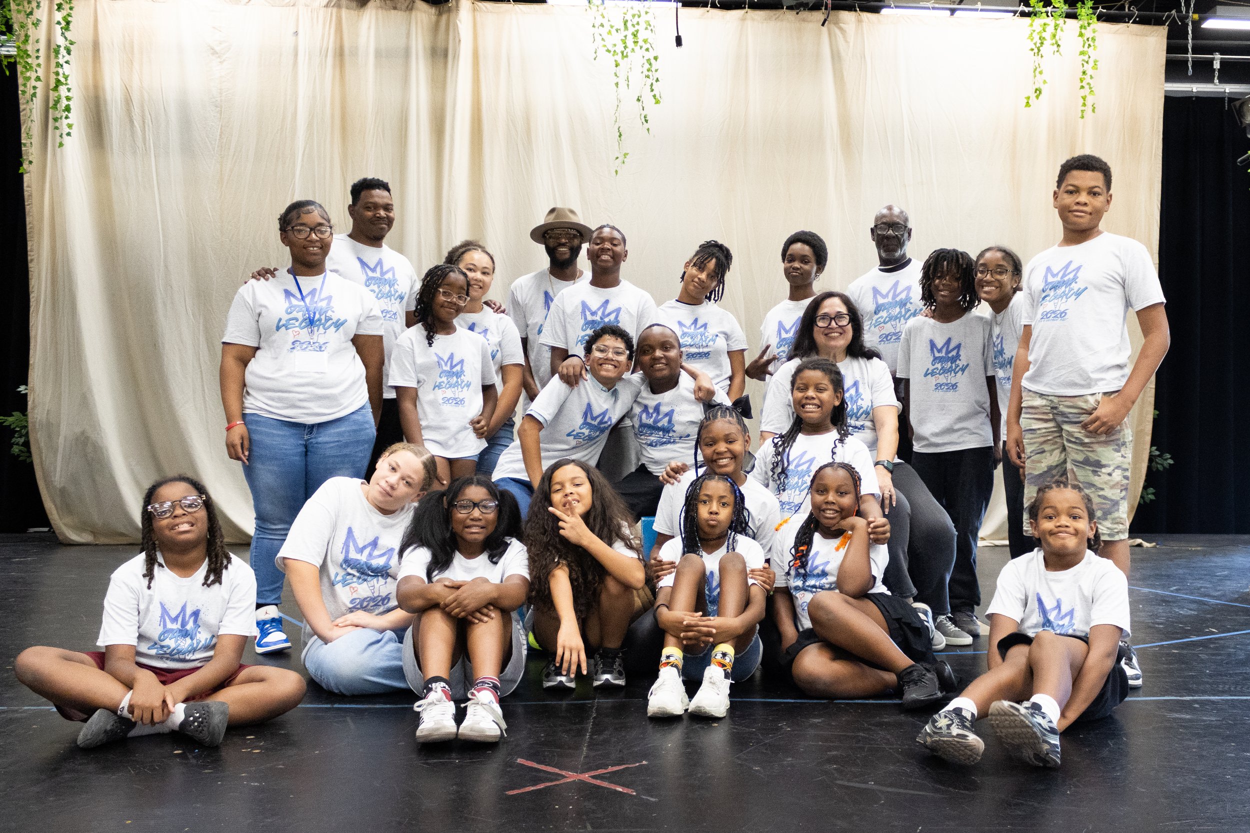 Group photo of the 2026 Summer Theatre Camp featuring youth students and staff wearing white camp shirts with blue print, standing on stage with a curtain backdrop.