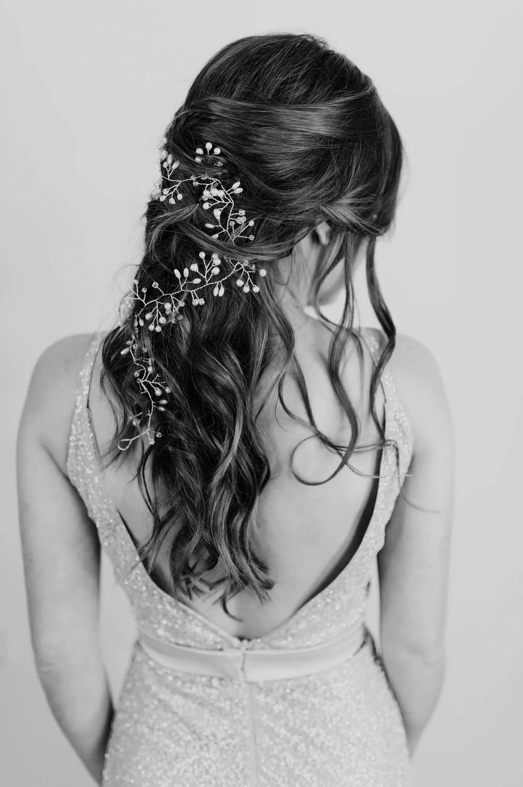 A woman with long, wavy hair styled in loose waves and adorned with delicate floral hair accessories. She is wearing a dress with a deep V neckline and thin straps, made of a sparkly, textured fabric. The image is in black and white, and the woman is looking downward with her face turned away from the camera.