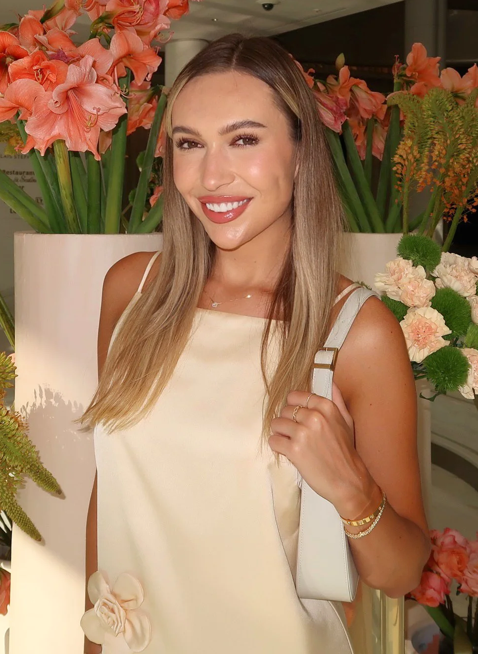 A woman with long blonde hair and a white dress standing in front of large pink and peach flowers.
