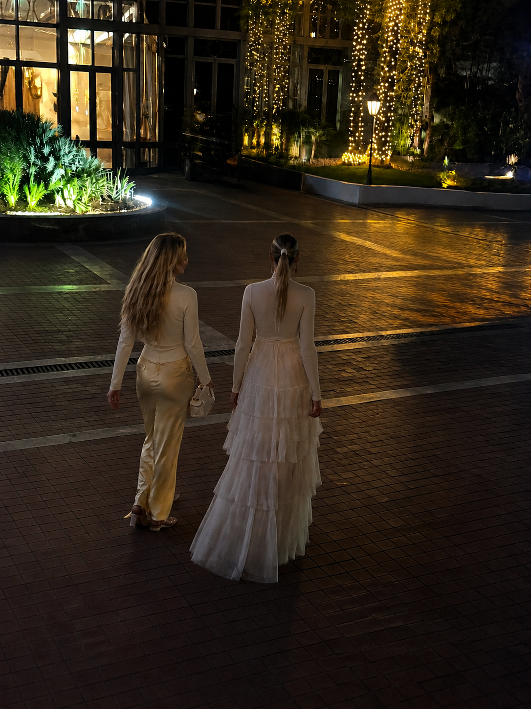 Two women dressed in elegant clothing walking on a wet tiled pavement at night, with a brightly lit greenhouse and decorated trees in the background.
