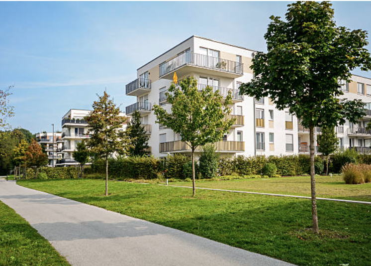 Modern apartment building with balconies surrounded by green trees and a grassy park with a walking path.