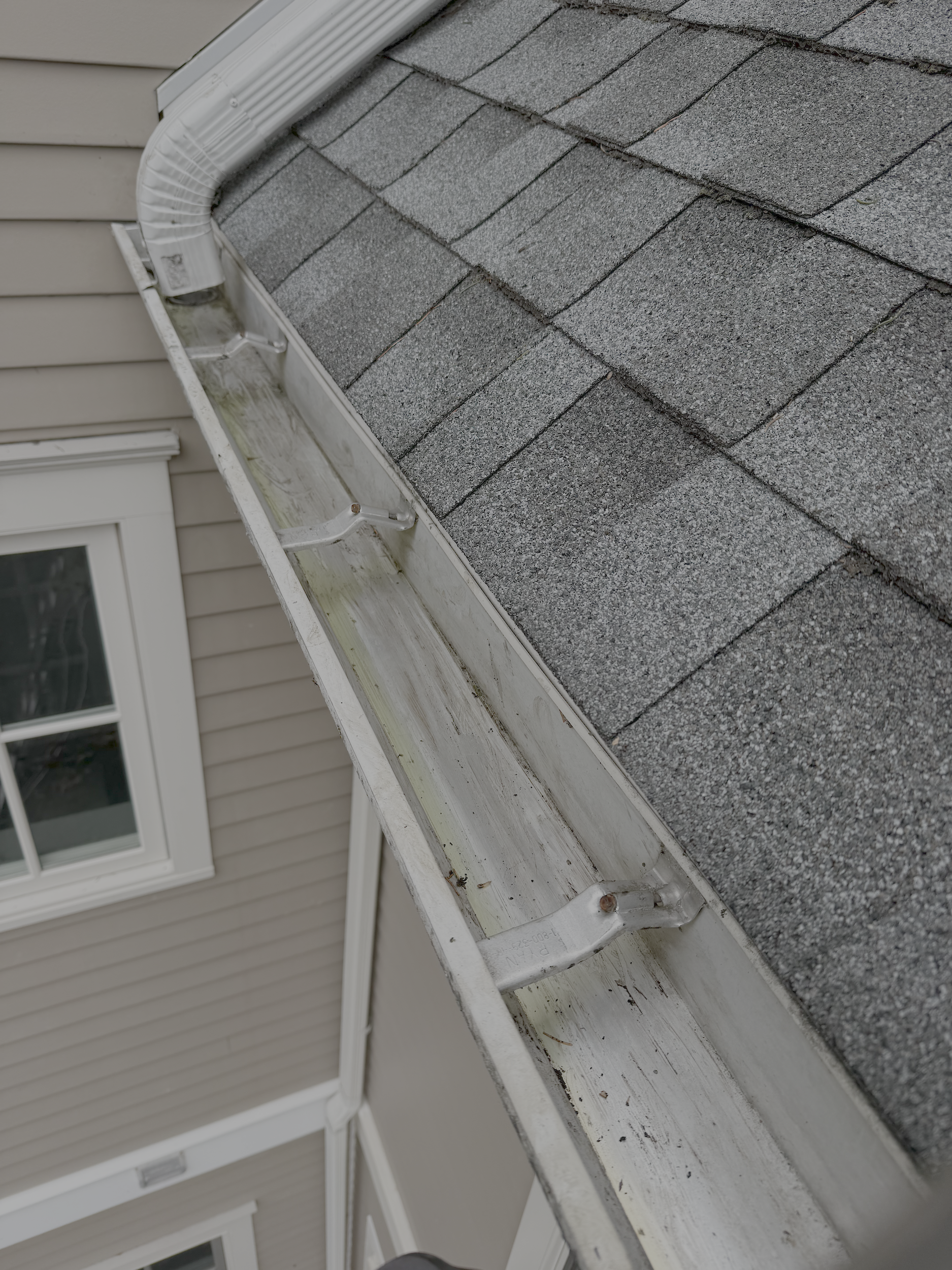Close-up view of a house's roof with gray shingles, part of the beige exterior wall, and a white gutter.
