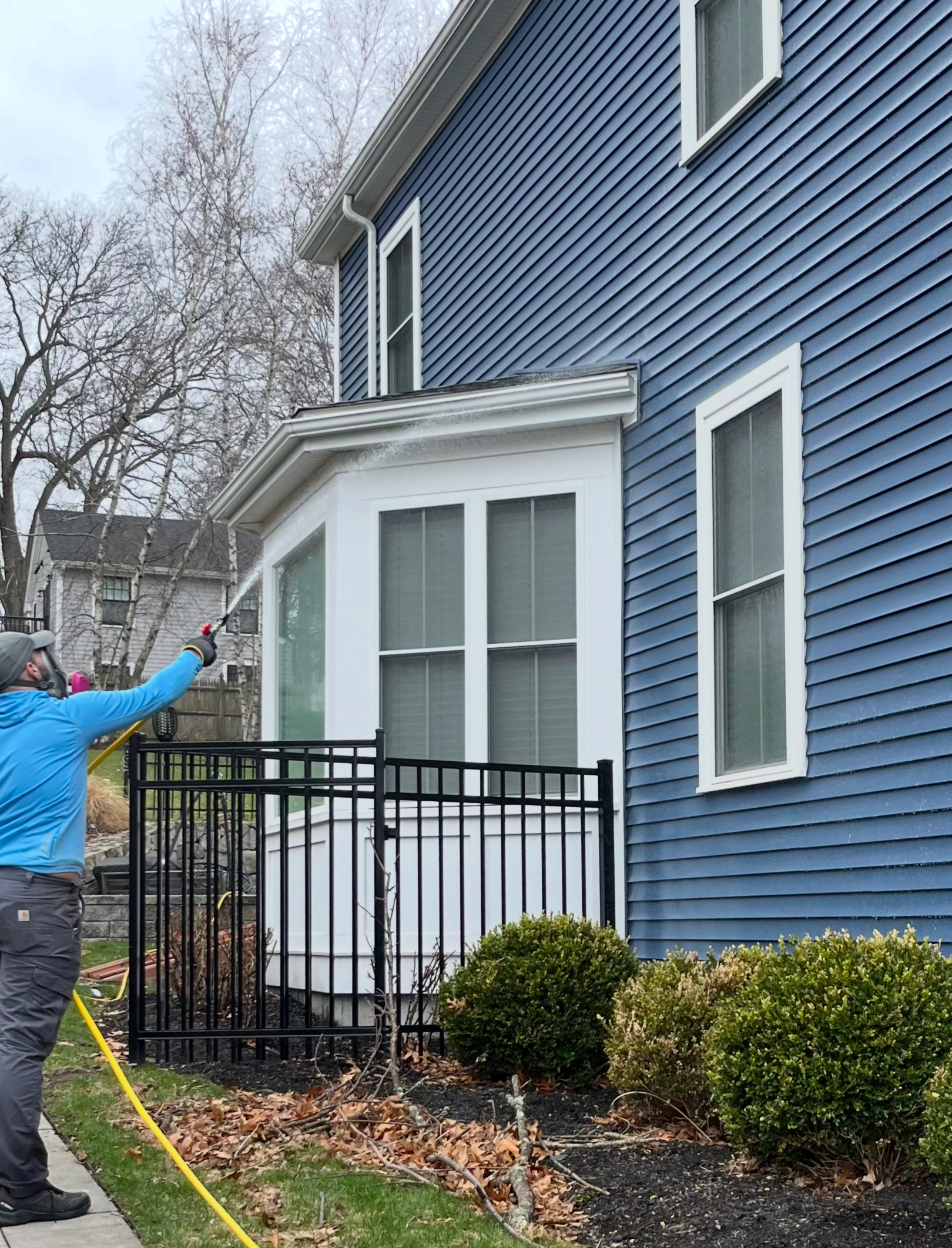 Jay Woodward, owner of Water Works, using a pressure washer to soft wash the exterior of a blue house with white trim, located near a black metal fence and bushes, in Hingham, MA.