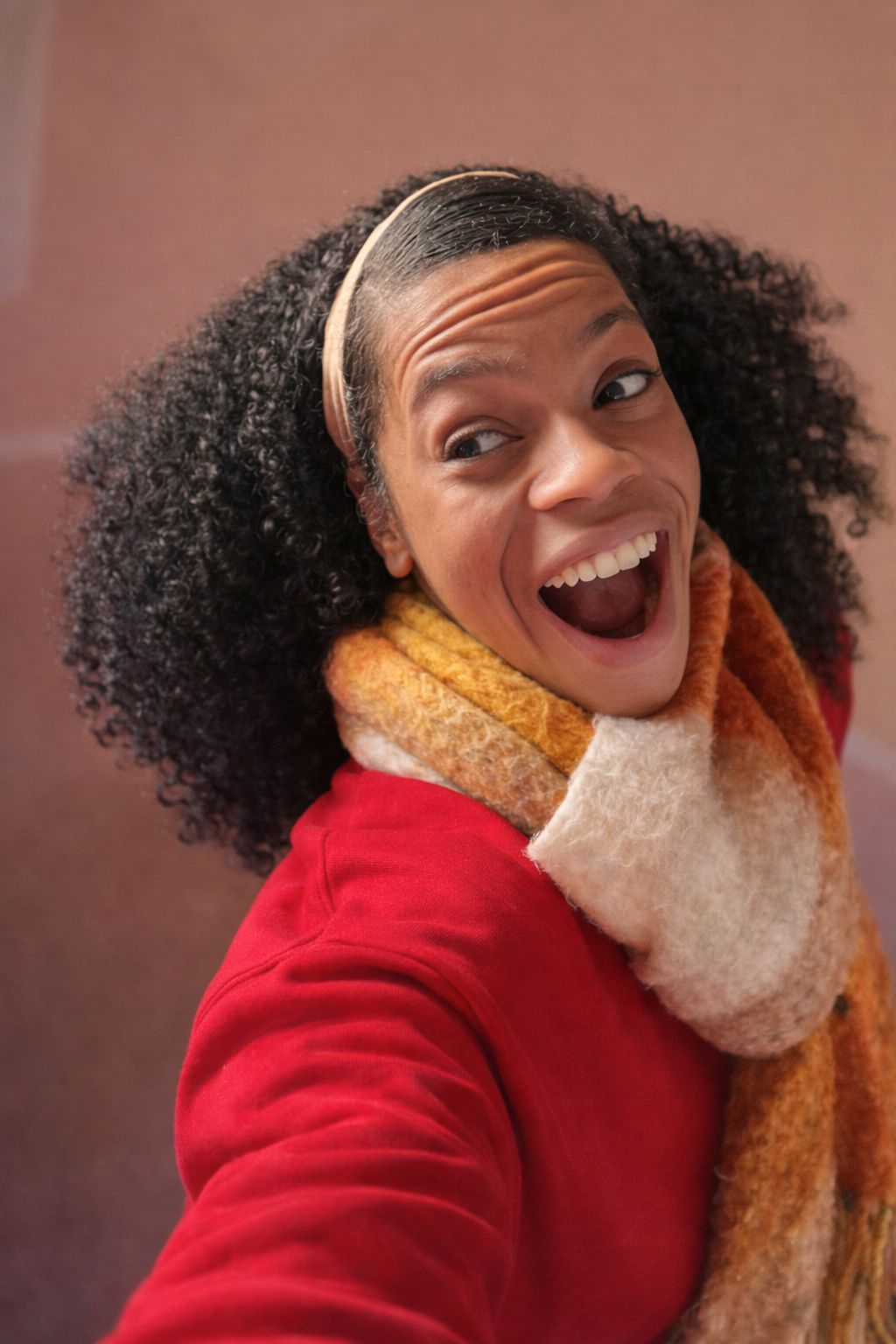 A woman with curly hair wearing a headband, orange and beige scarf, and red coat, smiling with her mouth open and looking off to the side.