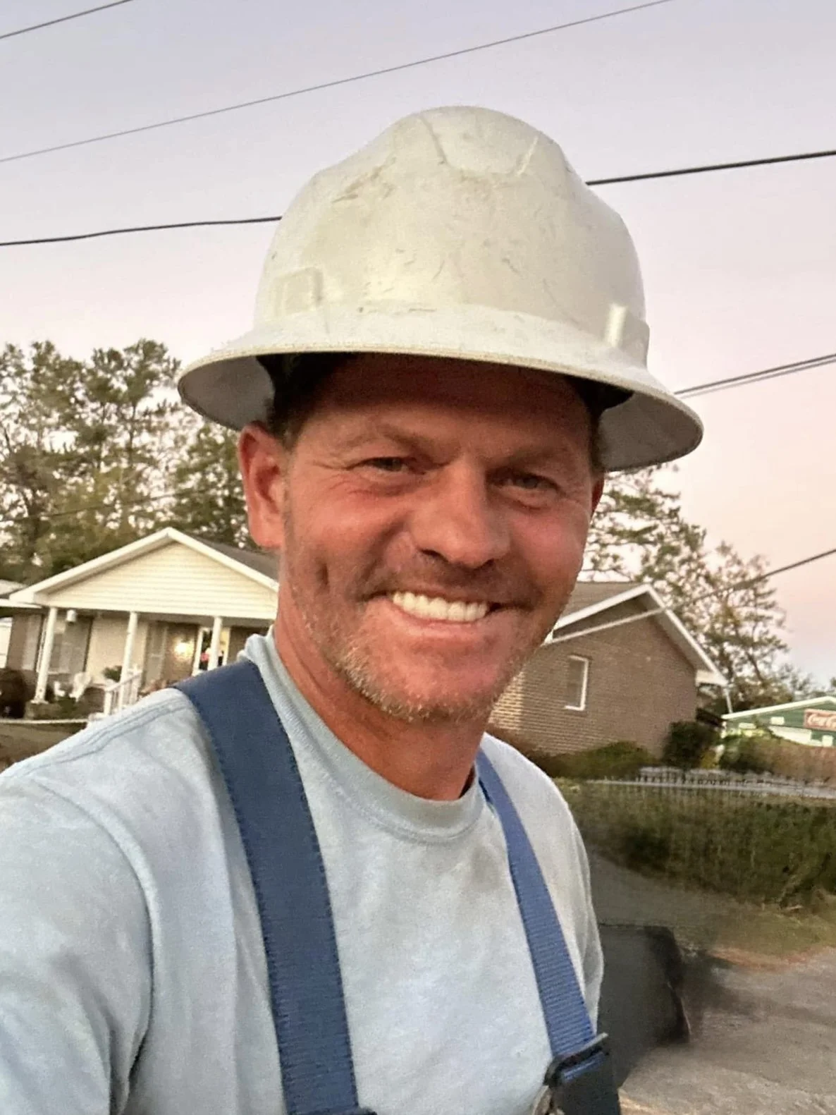 Smiling man wearing a white construction helmet and gray shirt outdoors with houses and trees in background.